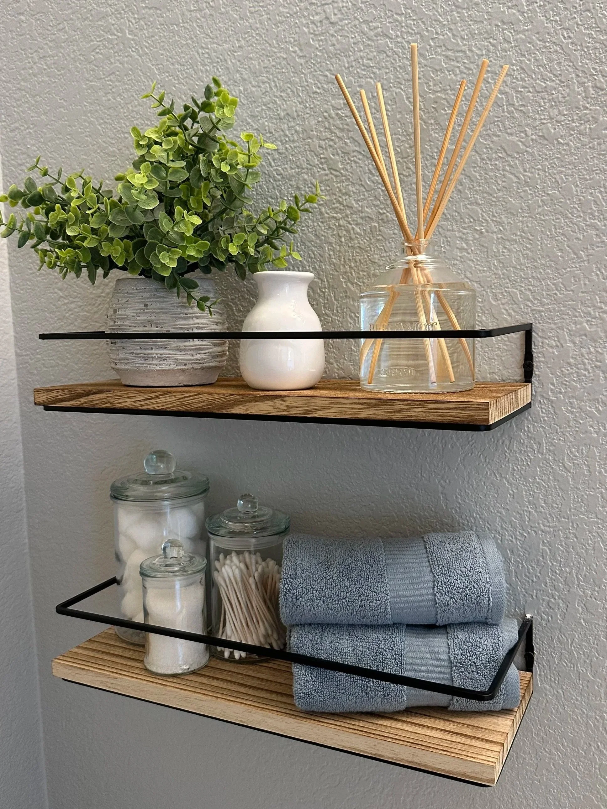 Decorative floating shelves with a potted plant, a small white vase, a diffuser, glass jars, cotton swabs, a towel, and a towel rod, against a textured wall.