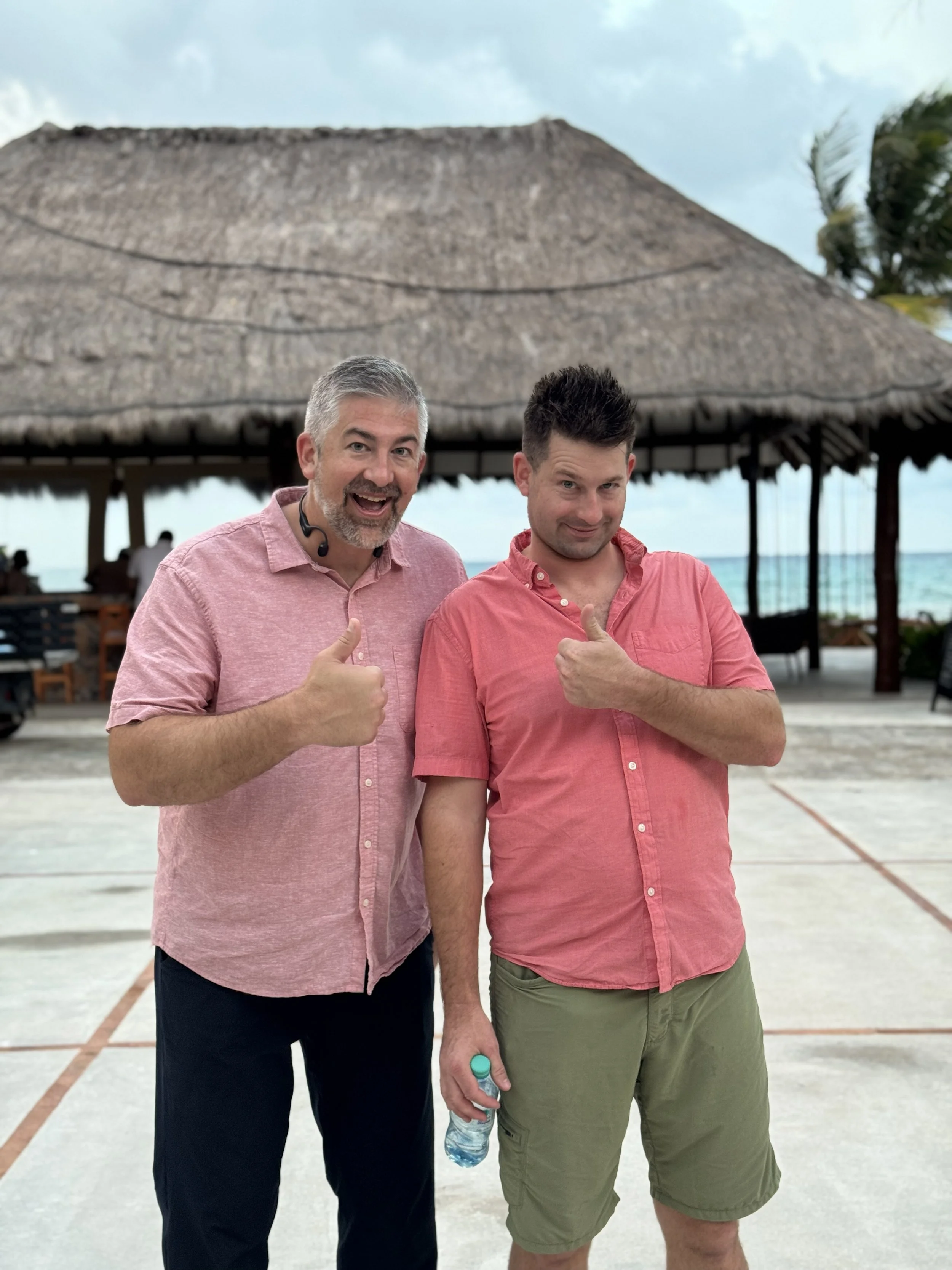 Two men standing outdoors at a beachside location, with a thatched roof structure in the background. The man on the left has gray hair and a beard, wearing a pink shirt and giving a thumbs-up. The man on the right has dark hair, wearing a pink shirt and shorts, holding a water bottle, and giving a thumbs-up. Both are smiling.
