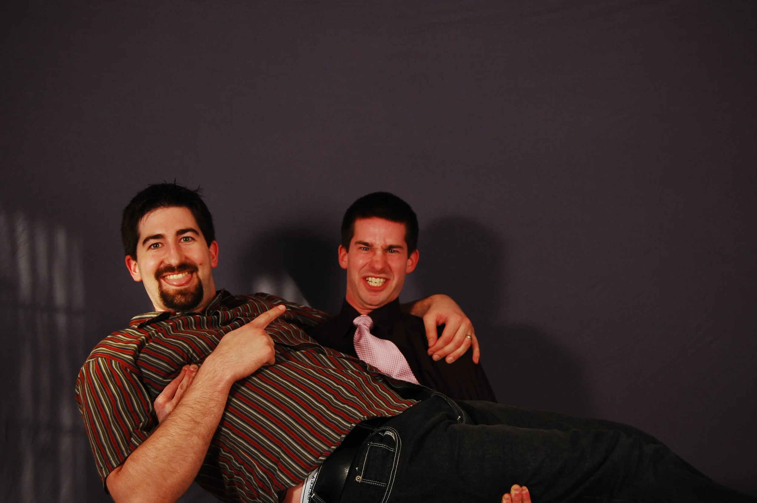 Two men smiling and posing together against a dark backdrop, one wearing a striped shirt and the other in a suit with a pink tie.