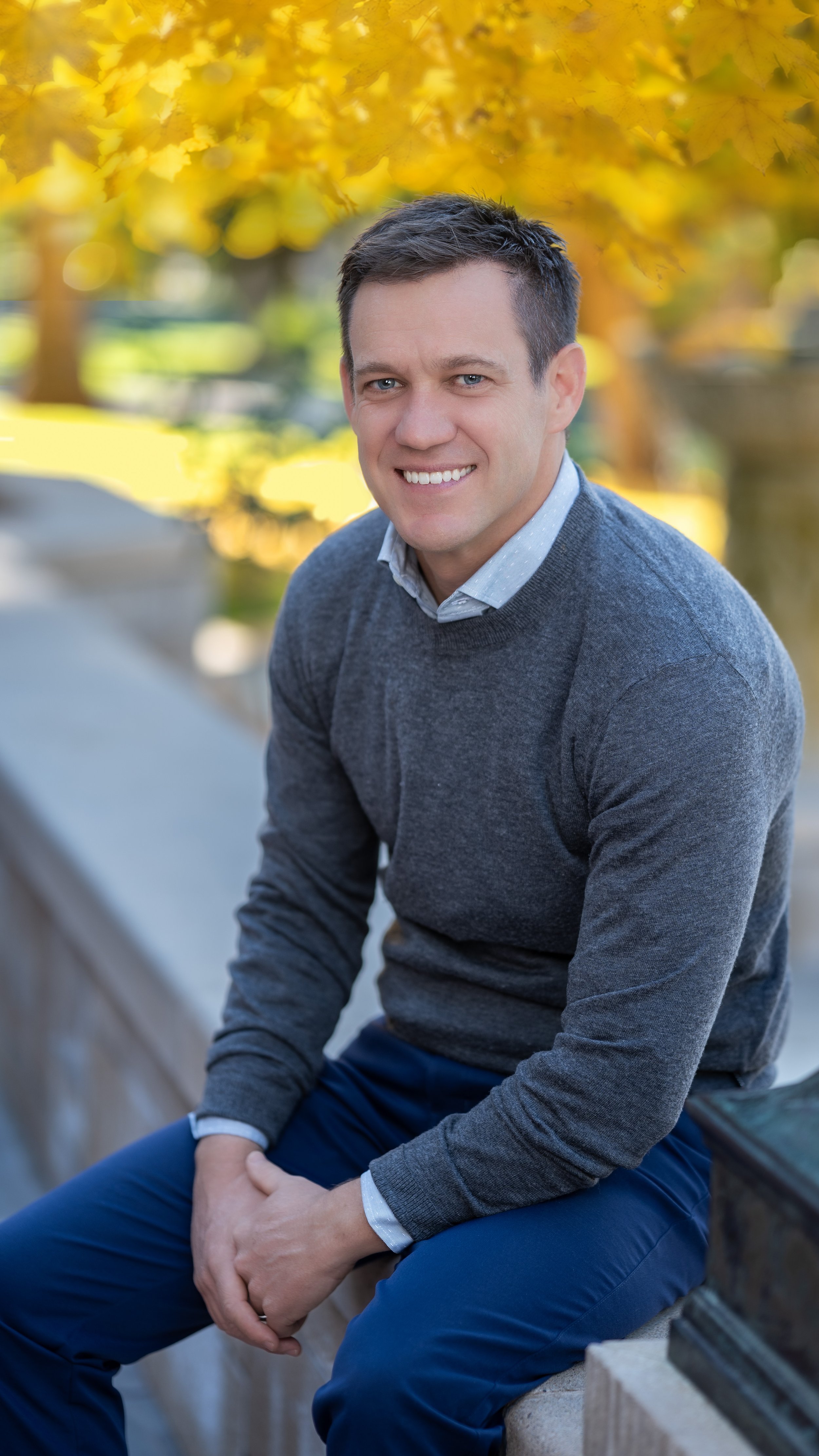 A man with short dark hair, smiling, wearing a light blue collared shirt under a dark gray sweater, and dark blue pants, sitting on a stone bench outdoors with yellow autumn leaves in the background.
