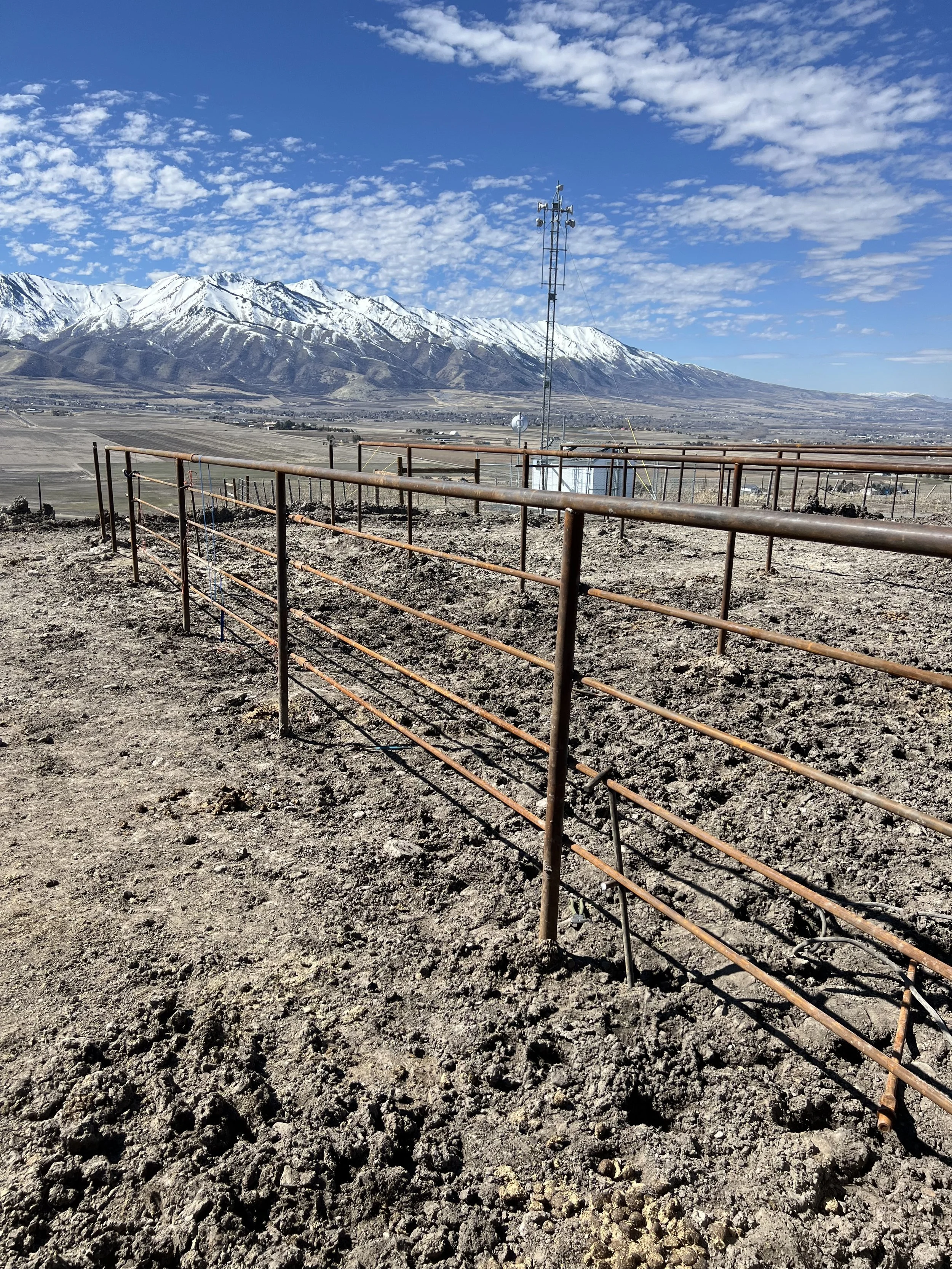 A barren dirt area with a metal fence in the foreground. In the background, snow-covered mountains under a partly cloudy blue sky.