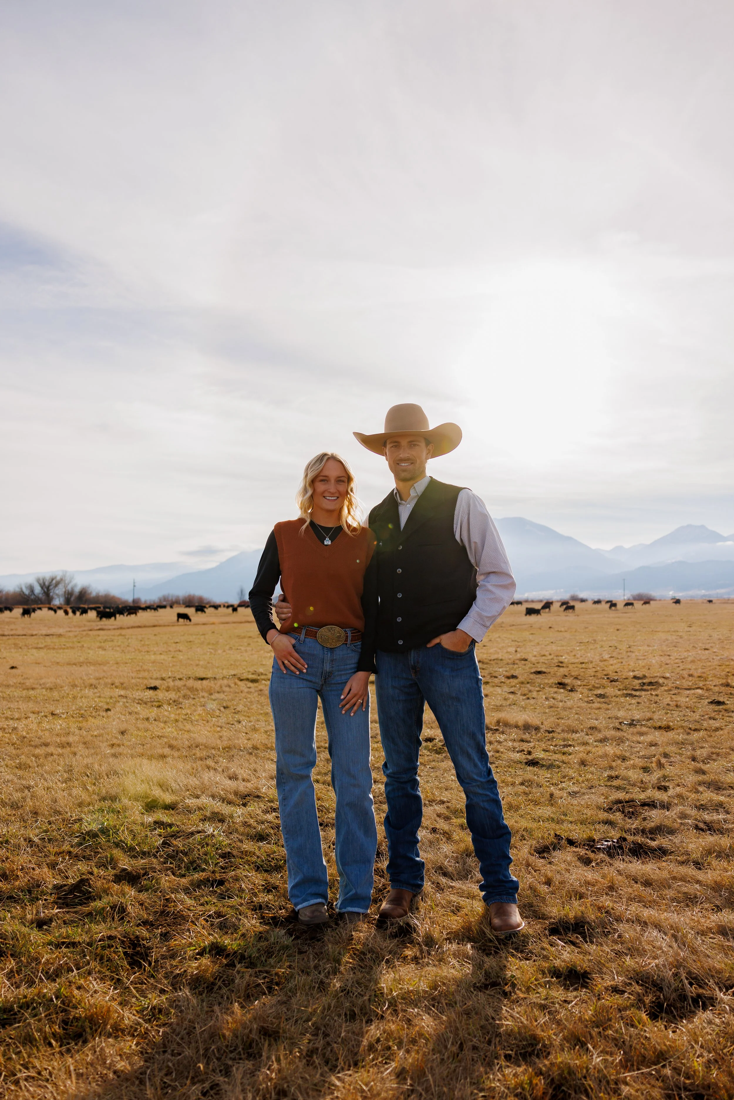 A smiling couple dressed in western attire standing in a vast, open field with horses grazing in the background and mountains in the distance under a partly cloudy sky.