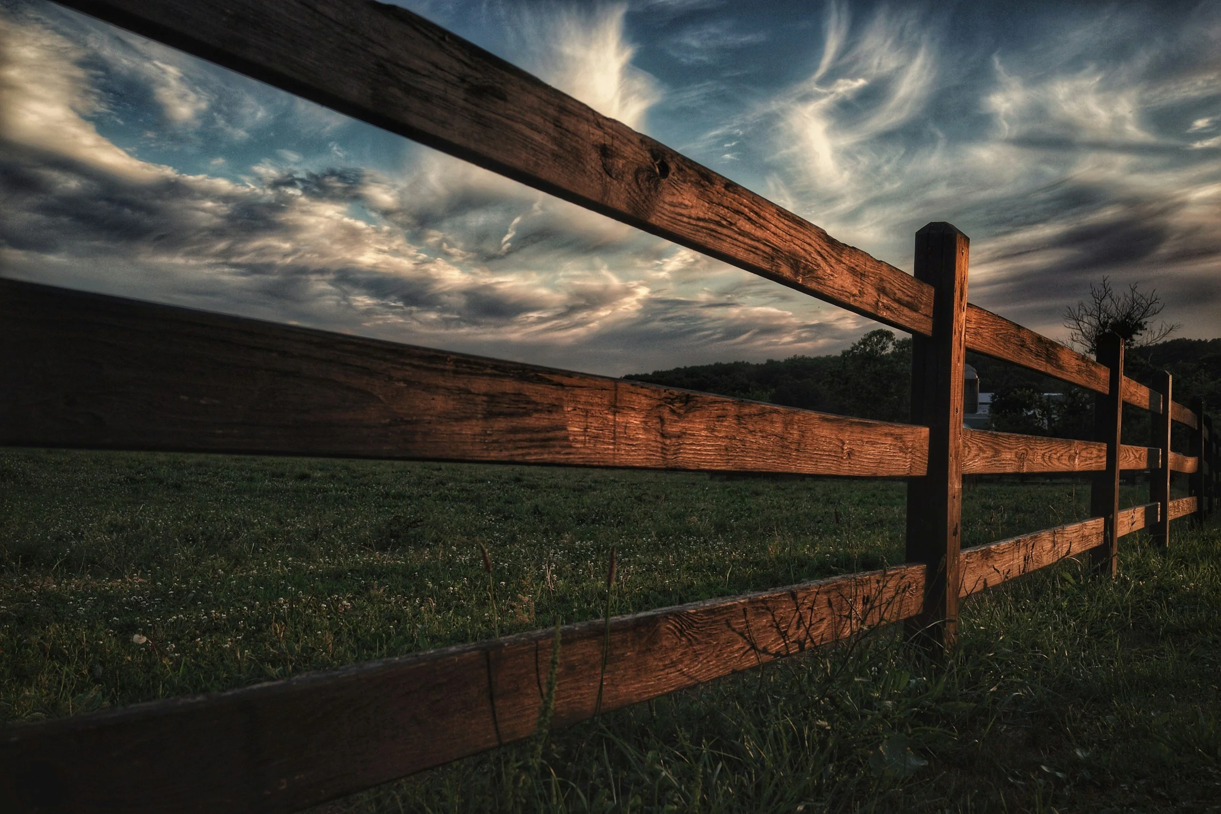 A wooden fence running through a grassy field against a cloudy sky during sunset.