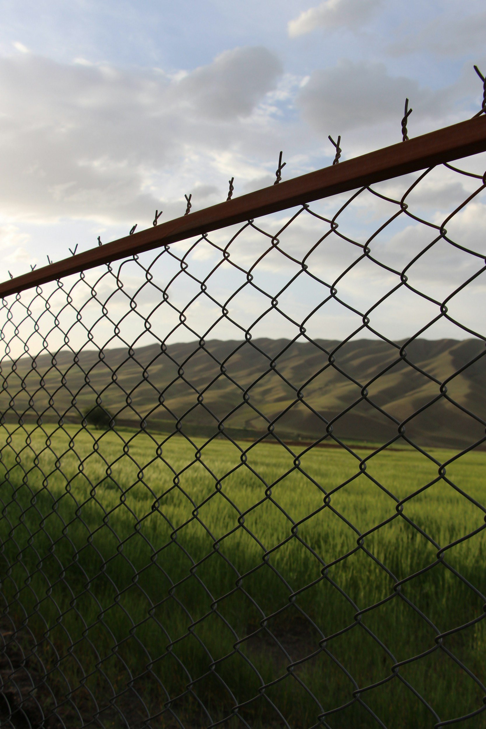 A chain-link fence with barbed wire on top, overlooking a landscape of green fields and rolling hills under a cloudy sky.