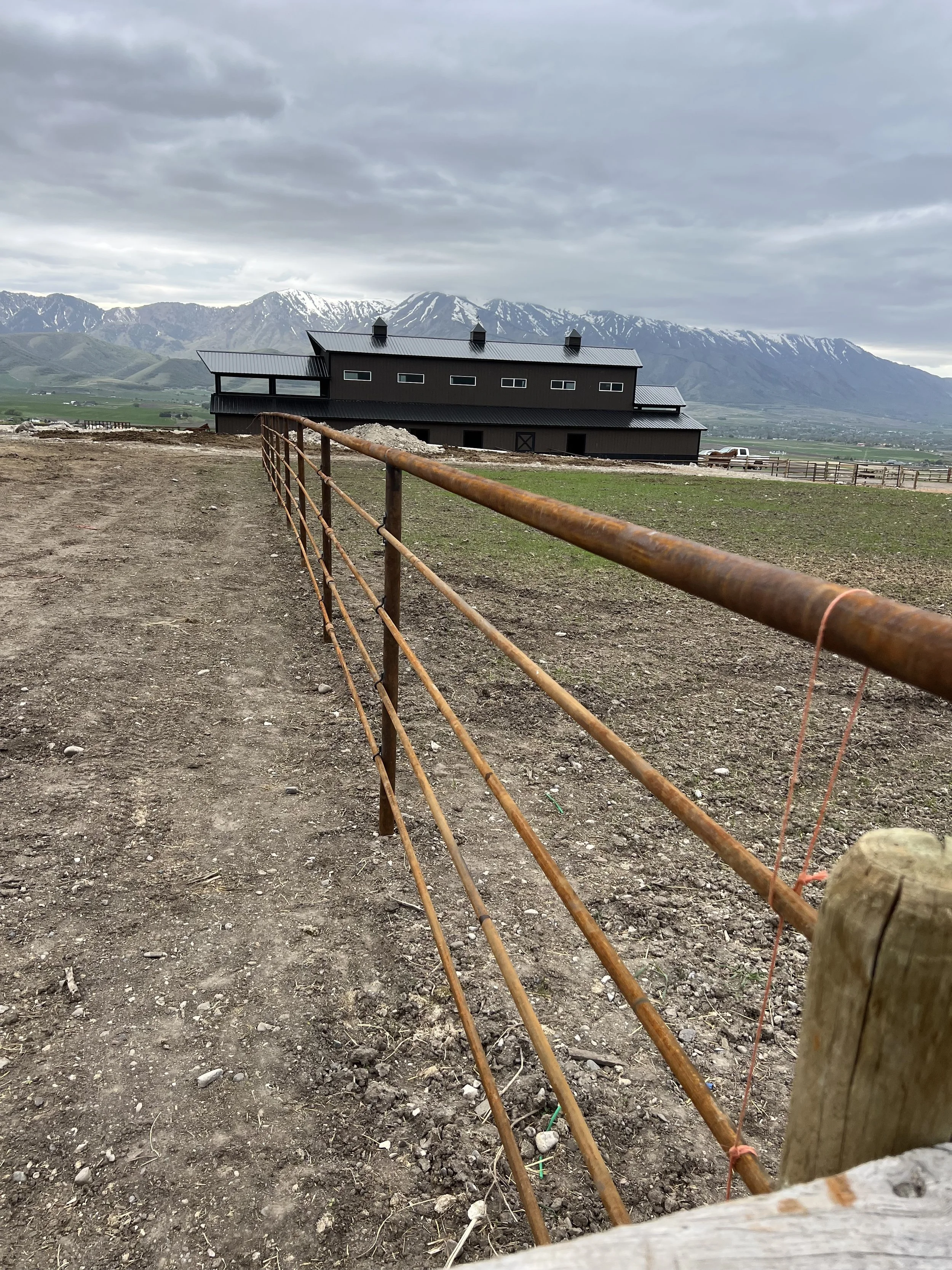 A fenced dirt path leading toward a black barn with a metal roof, set against a backdrop of mountains with snow-capped peaks under a cloudy sky.