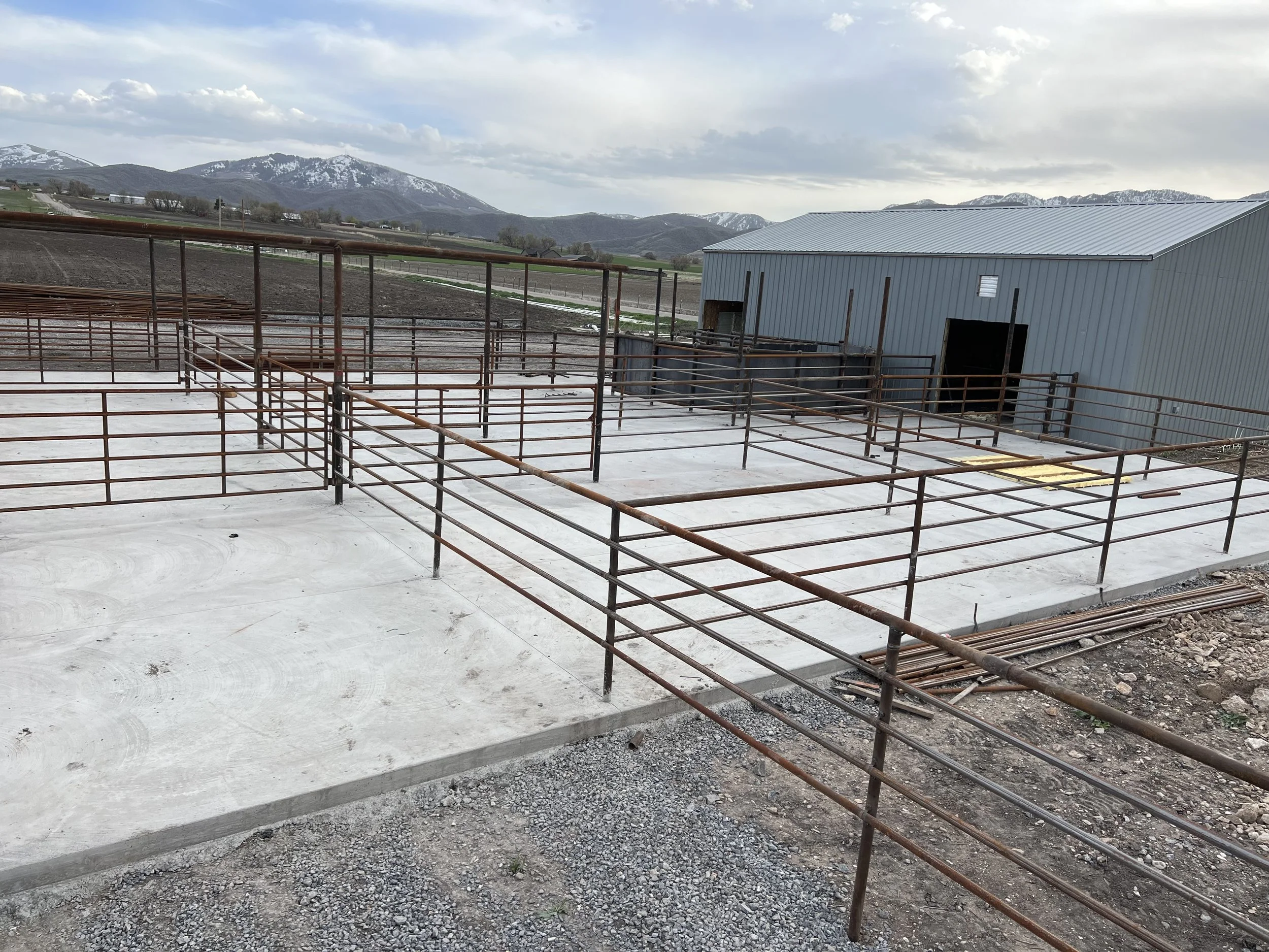 A construction site with a concrete foundation and metal framing, set against a mountainous landscape with snow-capped peaks and partly cloudy sky.