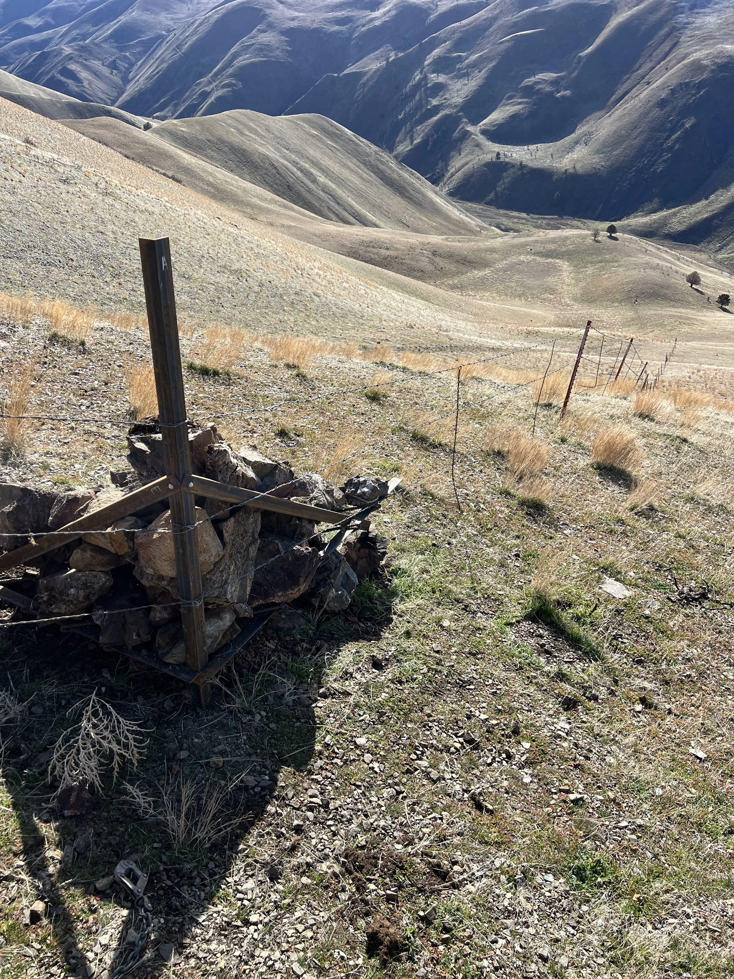A hillside with sparse grass and rocky terrain in a mountainous area, a barbed wire fence runs along the slope, and there is a metal post in the foreground.