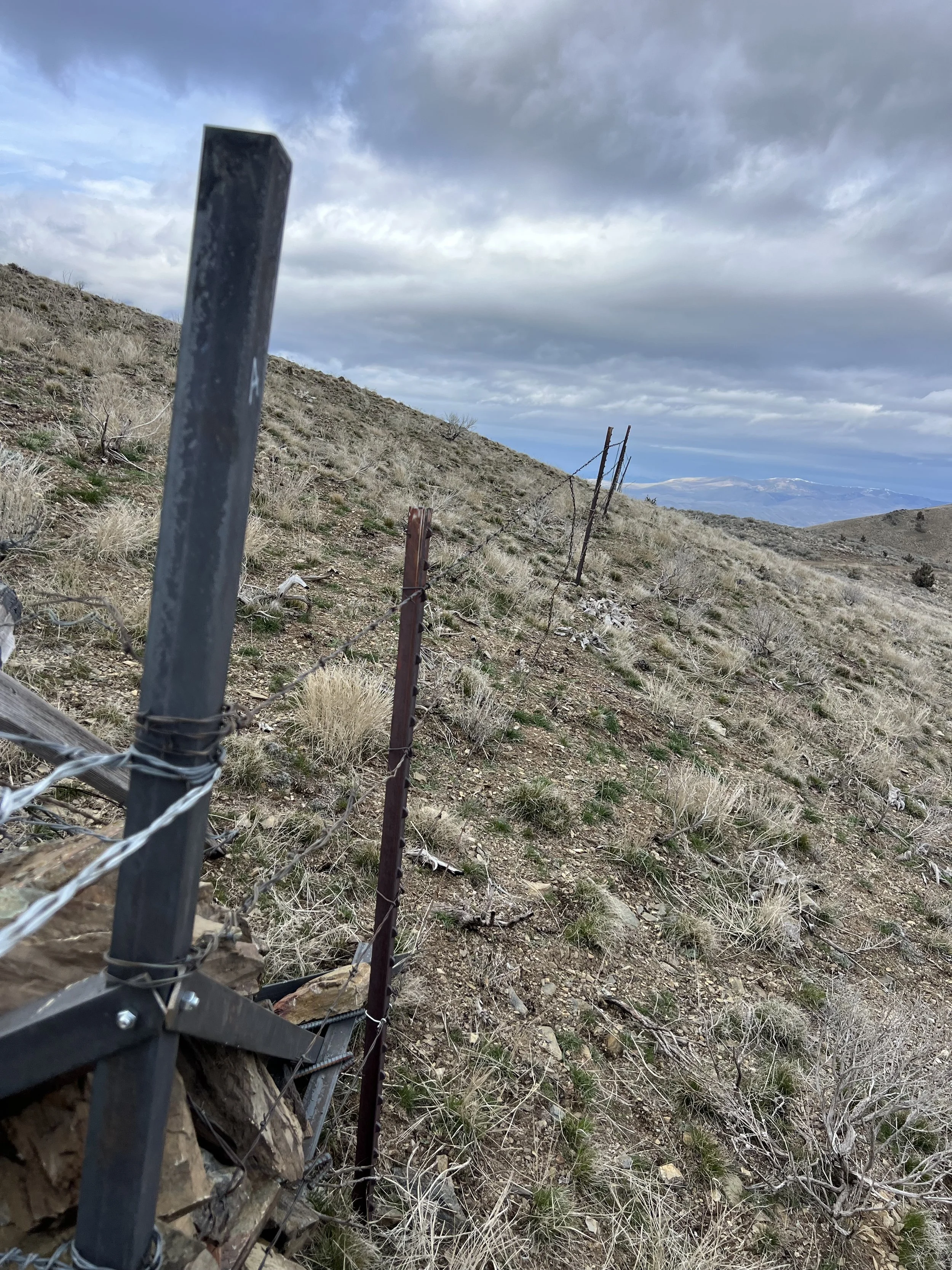 A hillside with a barbed wire fence that is broken and leaning, with cloudy sky and mountains in the background.