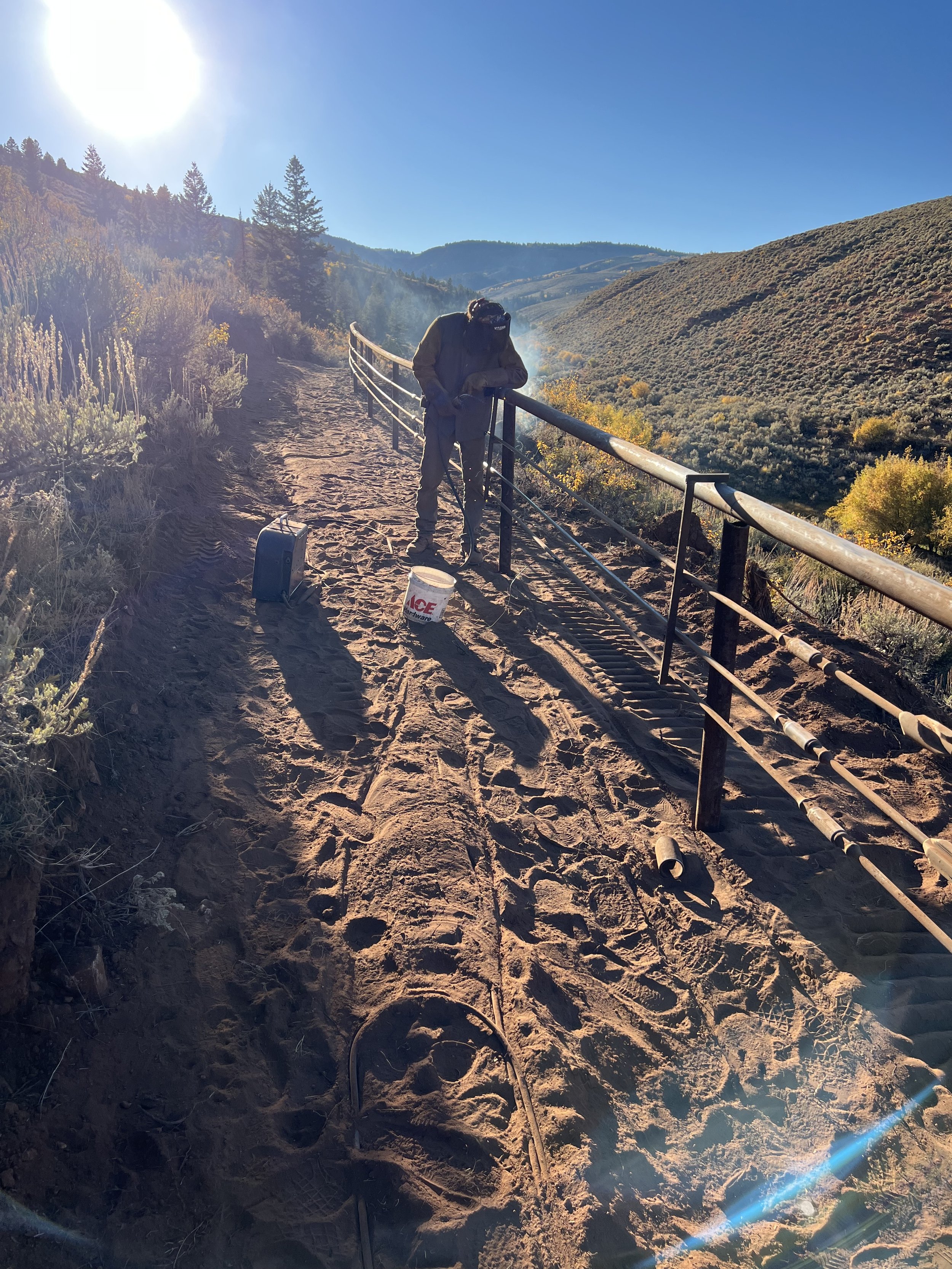 A person wearing a dark jacket and cap is working on a hillside overlooking a valley with trees and mountains under a bright blue sky, using tools on the ground near a bucket and a small black case.