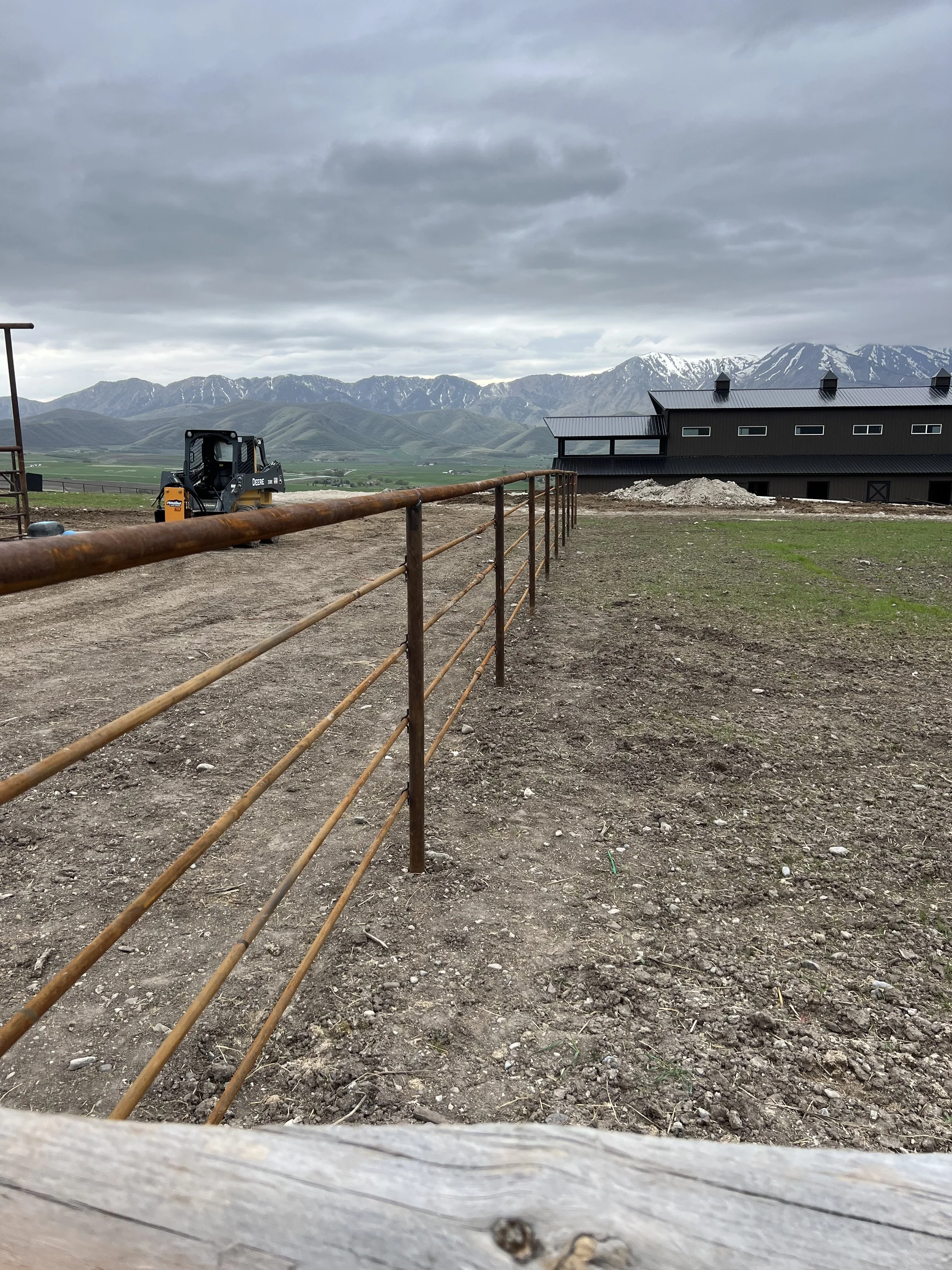 A rural scene with a black barn, a small tree, a rusty metal fence, a yellow skid steer loader, and snow-capped mountains in the background under cloudy skies.