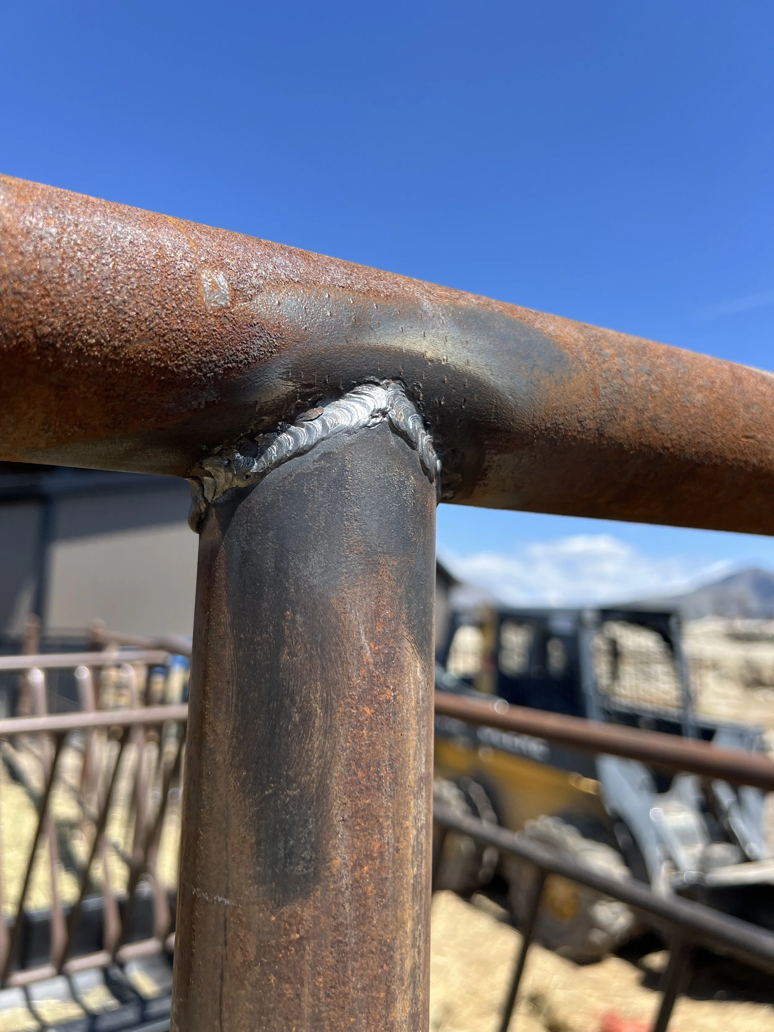 Close-up of a rusty metal pipe with a welded joint, against a background of blurred outdoor furniture and a blue sky.