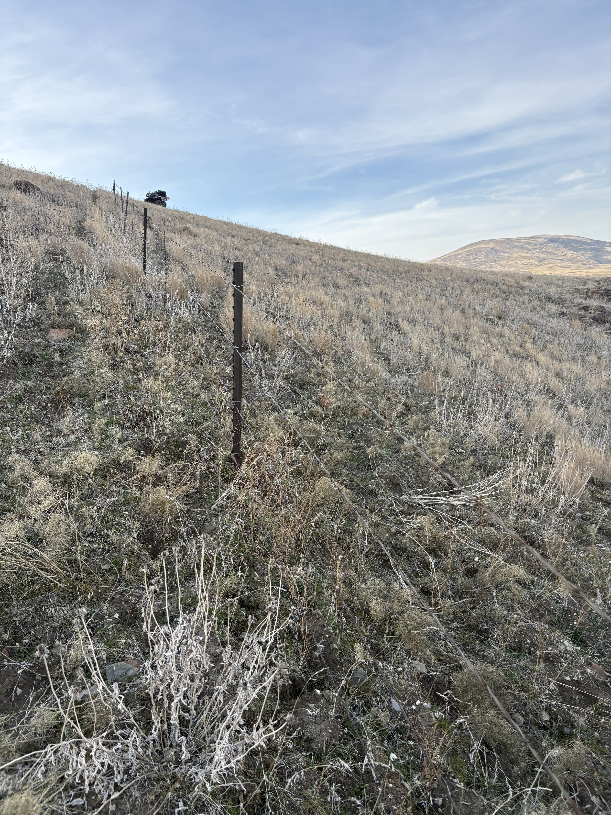 A dry, grassy hillside with sparse vegetation, a barbed wire fence running down the slope, and a black pickup truck parked at the top of the hill against a partly cloudy sky.