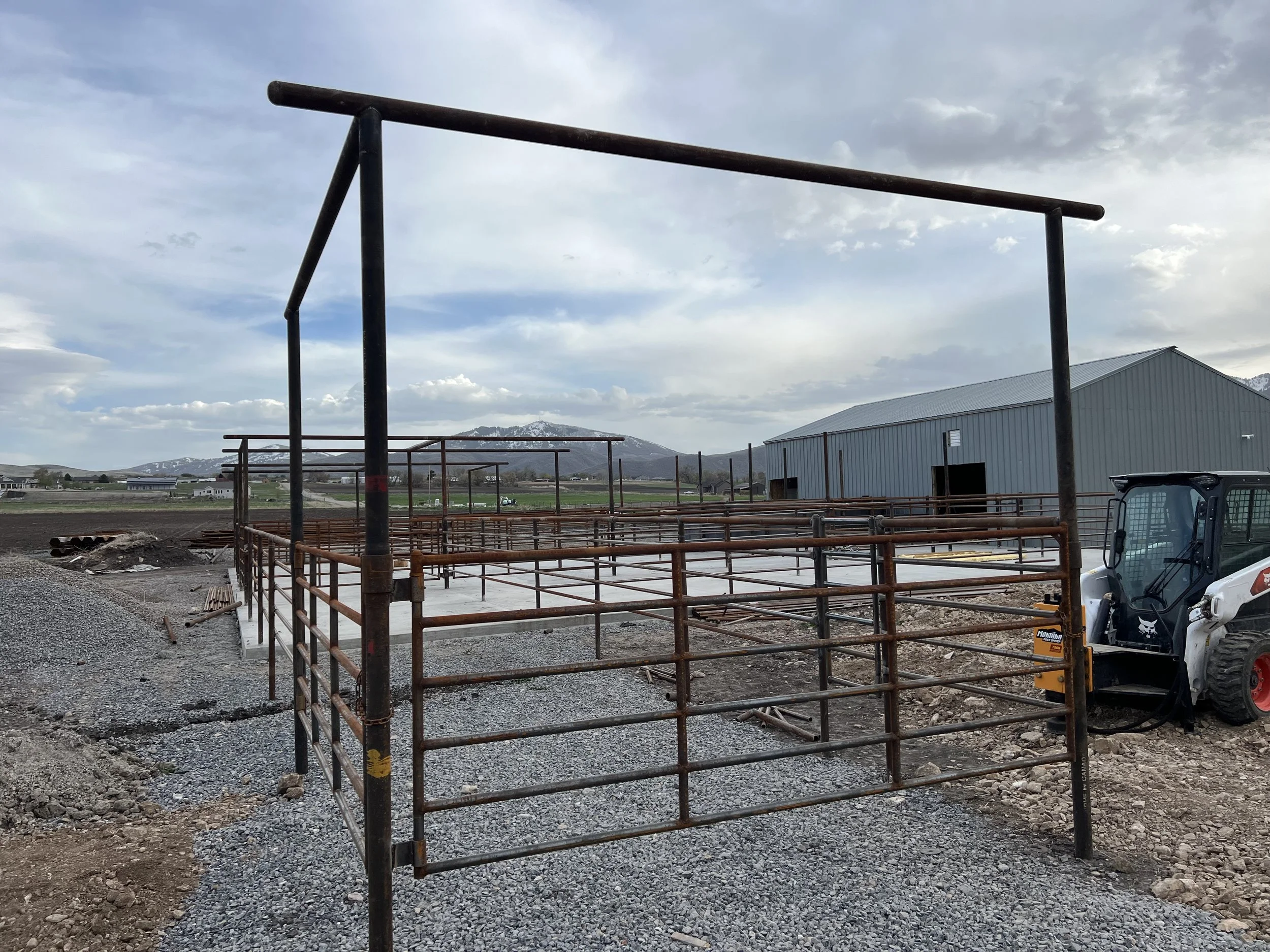 Construction site with metal frame structures and a small excavator in an open area, with a warehouse and mountain range in the background under a cloudy sky.