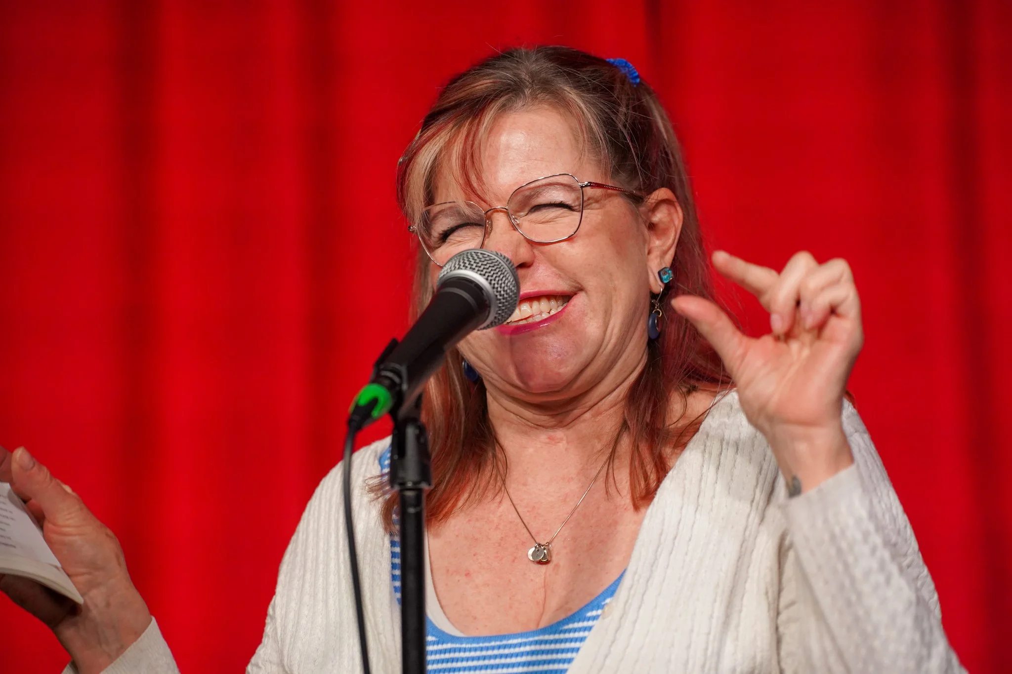 A woman with glasses and red hair, smiling and laughing near a microphone, with a red curtain background.