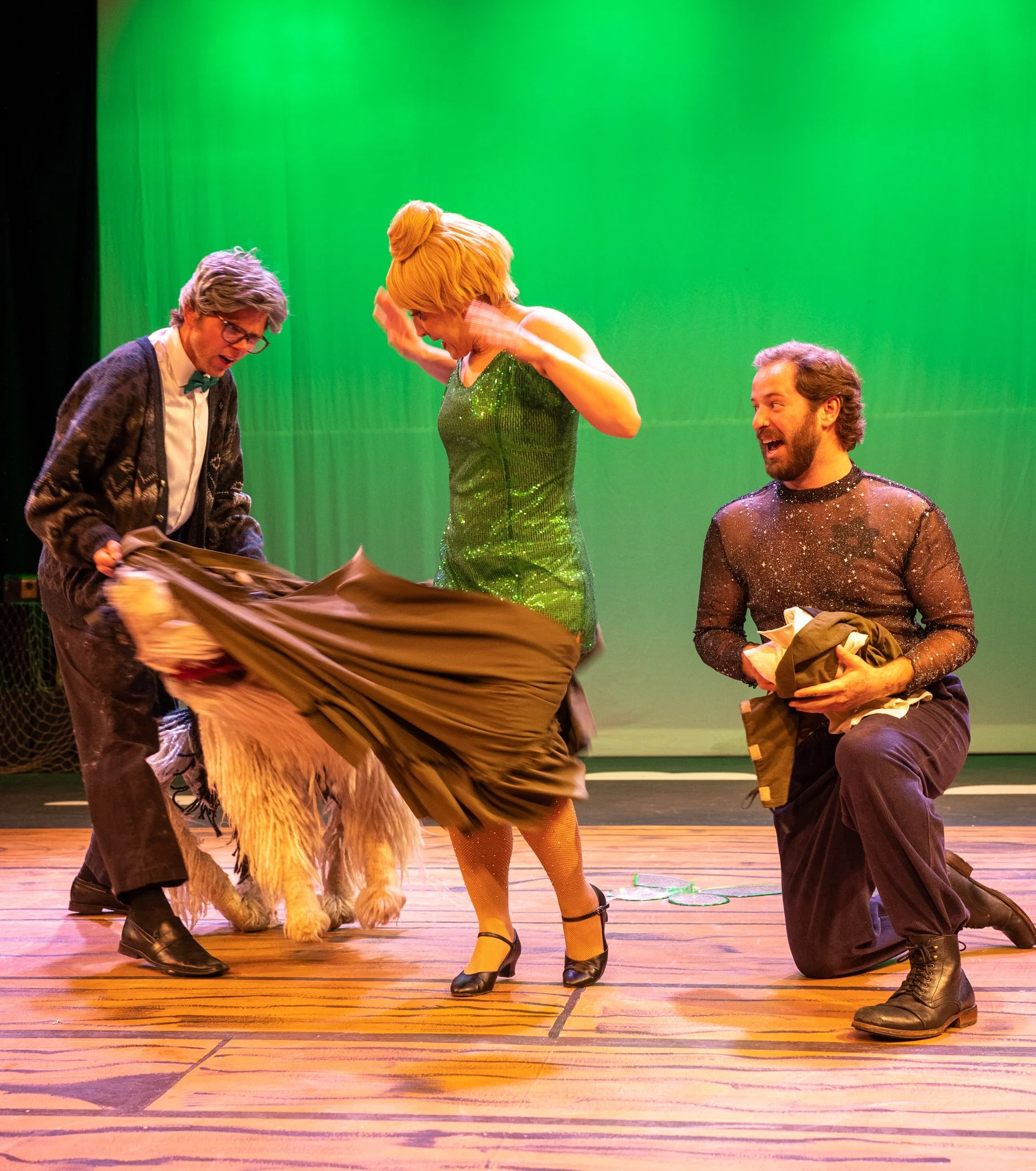 Four performers on stage with a green background. One is holding a large brown cloth, a woman is in a short, green, sequined dress and high heels, one man is kneeling and smiling, and another man with glasses and a cardigan is looking down. The scene
