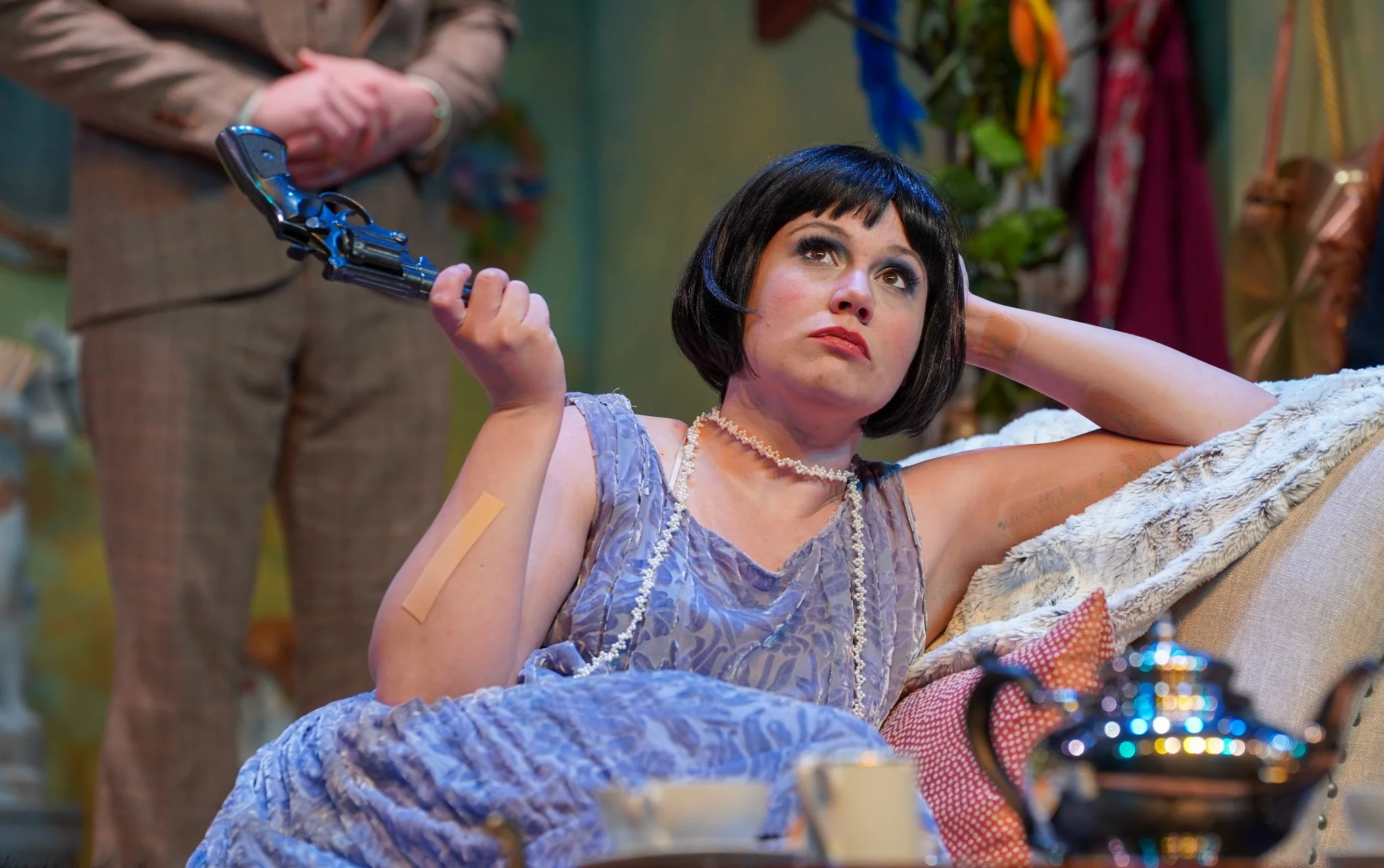 A woman with black hair styled in a bob wearing a purple dress adorned with pearls, lying on a plush chair, holding a lever of a machine, with a concerned expression, in a colorful room with various hanging fabrics and objects.