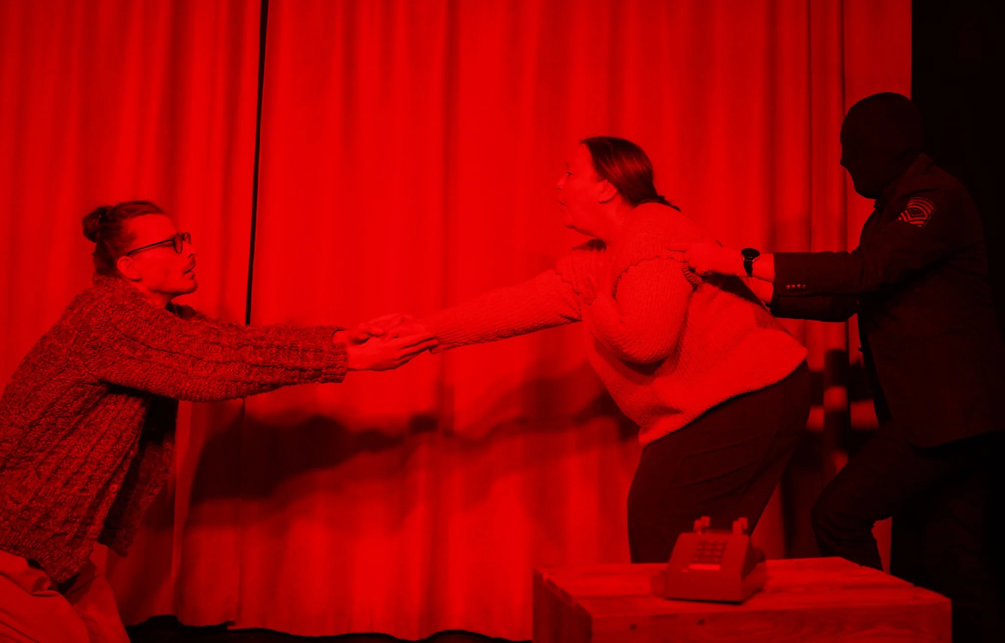 An emotional reunion scene with two women and a man in a dark uniform, possibly a police officer, with a red background curtain. One woman is on her knees with her hands extended, and the other woman is leaning forward, holding her hands. The man is 