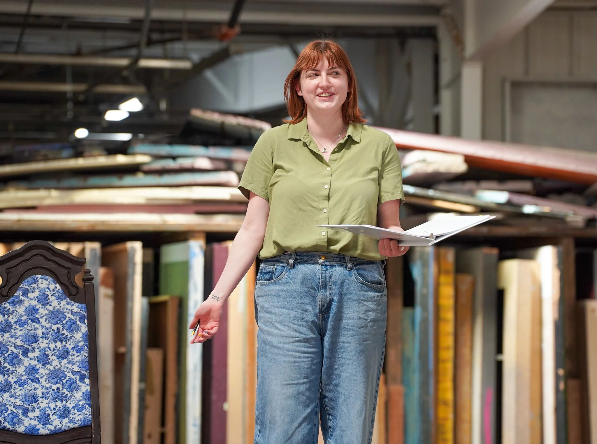 A red-haired woman in a green shirt and blue jeans holding papers and pencils, standing in a warehouse with stacks of chairs and shelves.