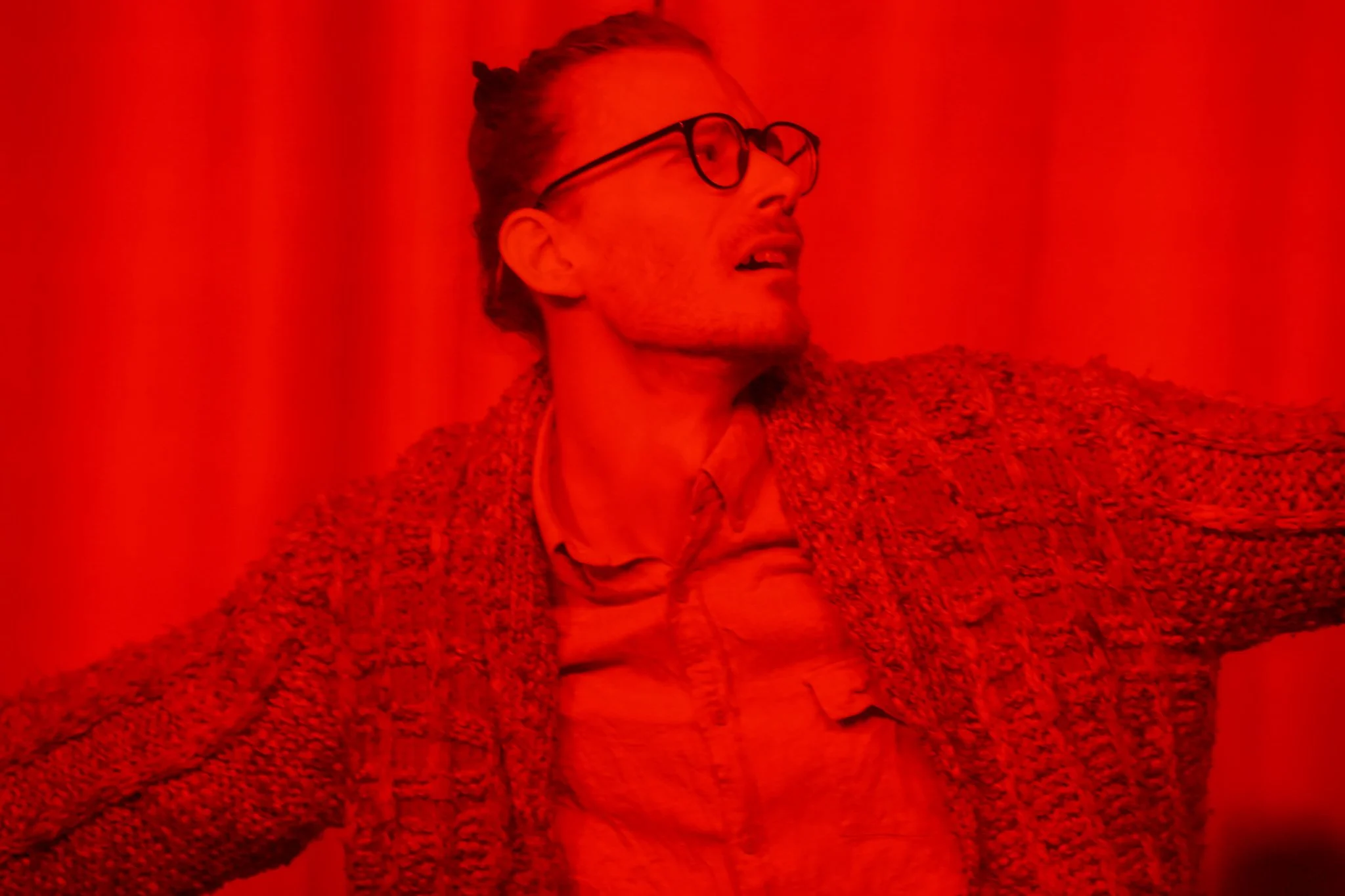 A person with glasses and light-colored hair tied back, wearing a textured jacket, leaning back against a bright red background.