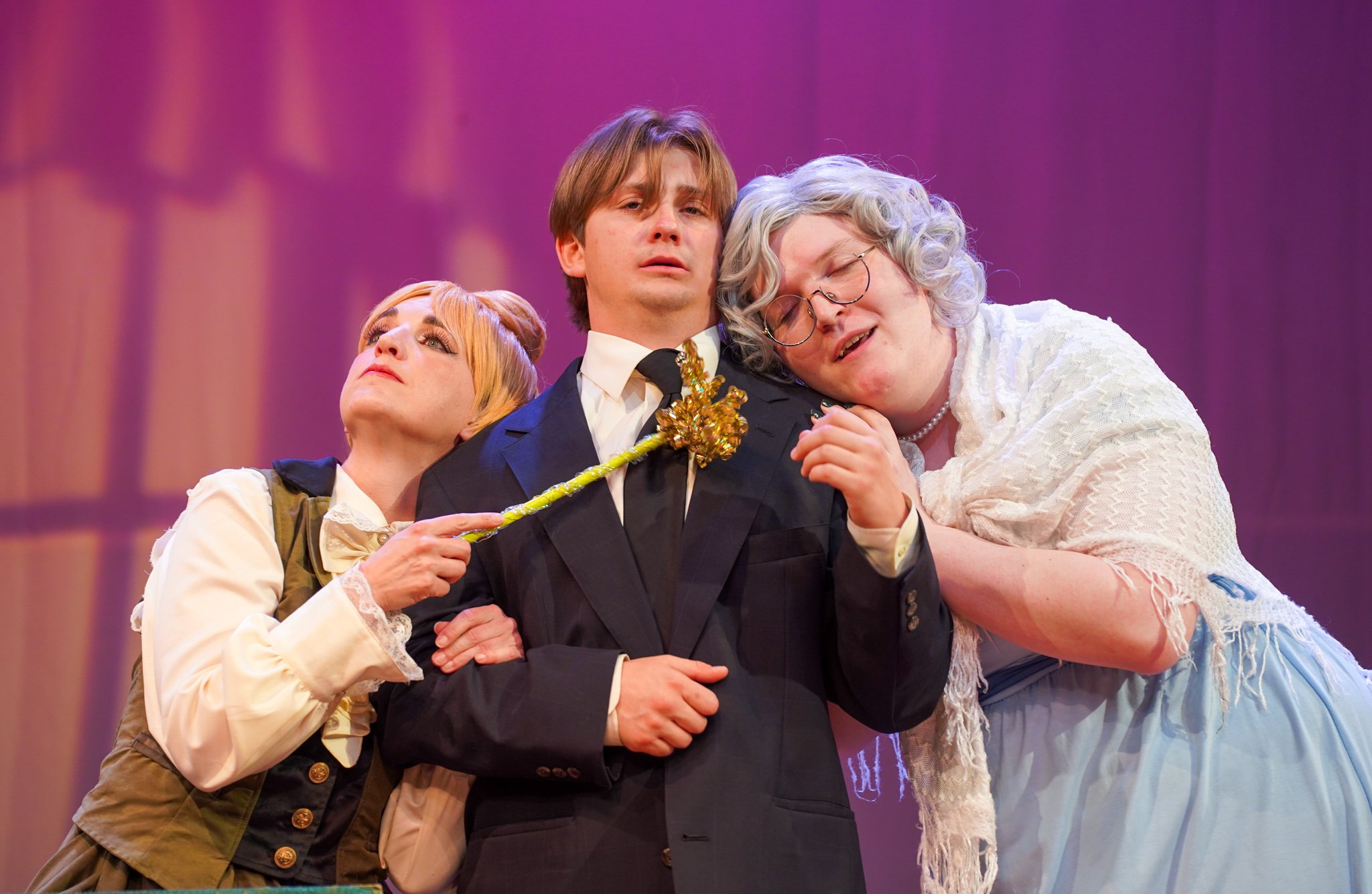 Three people engaged in a theatrical scene with a pink and purple background. The central figure, a man in a formal suit, is being hugged by a woman with curly gray hair wearing glasses and a white shawl. Another woman with reddish hair and vintage-s