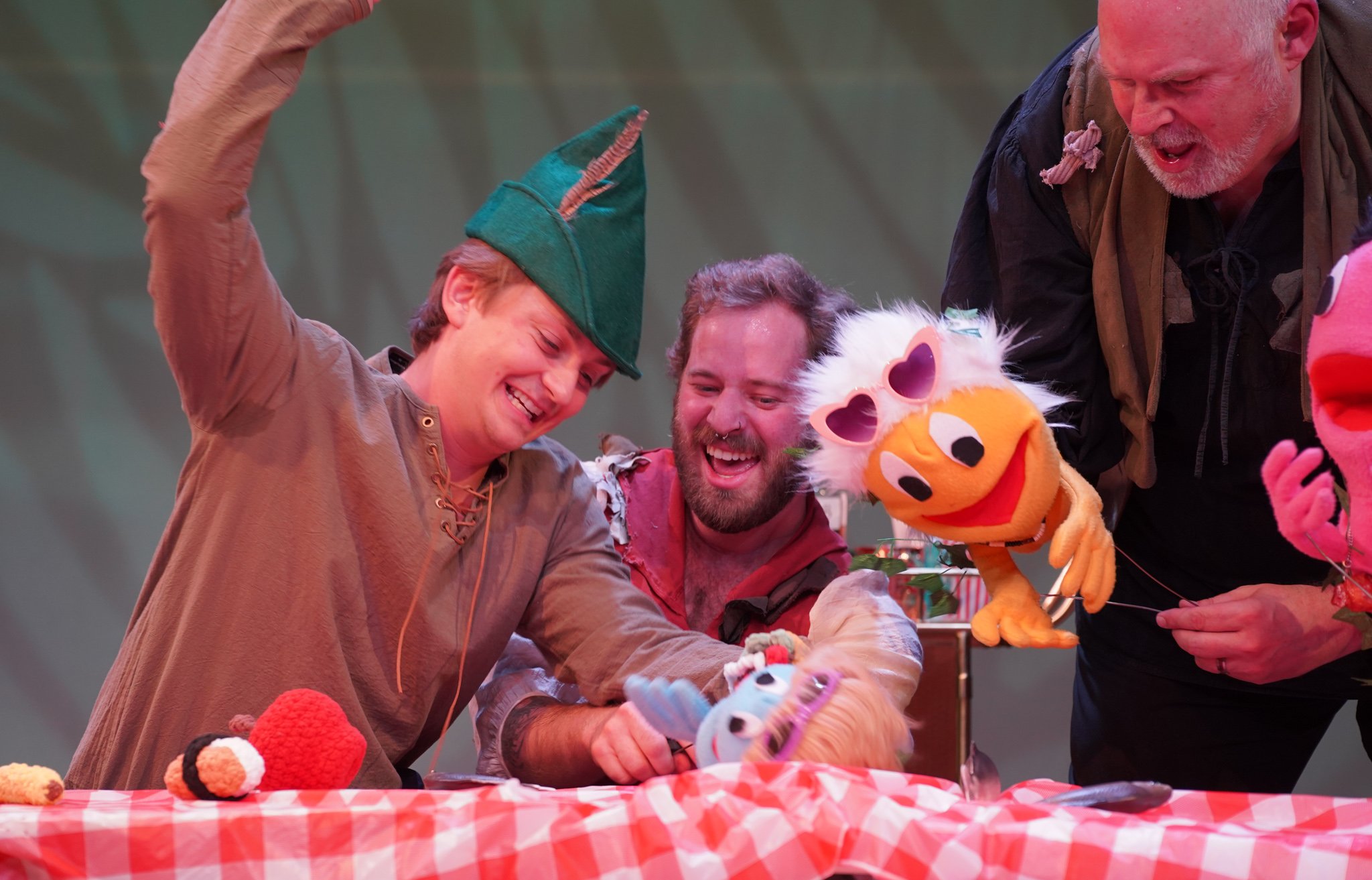 A group of people at a table with a red and white checkered tablecloth, enjoying a puppetry or craft activity involving puppet characters such as a yellow lion with pink sunglasses and other colorful puppets.