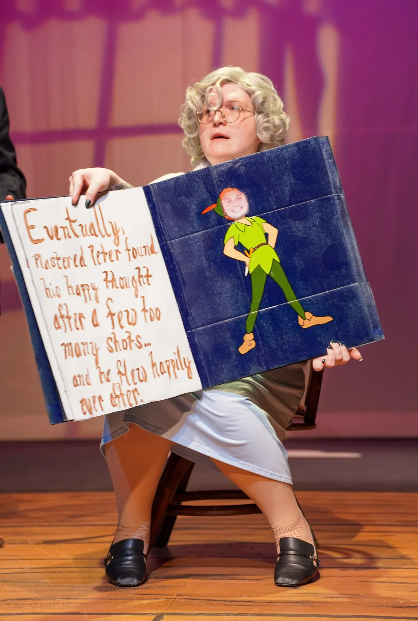 A person with curly blonde hair and glasses sitting on a chair holding a poster. The poster has an illustration of a cartoon character, Peter Pan, with the face of the person, and text that reads: "Eventually, I Plastered Peter Found his happy though