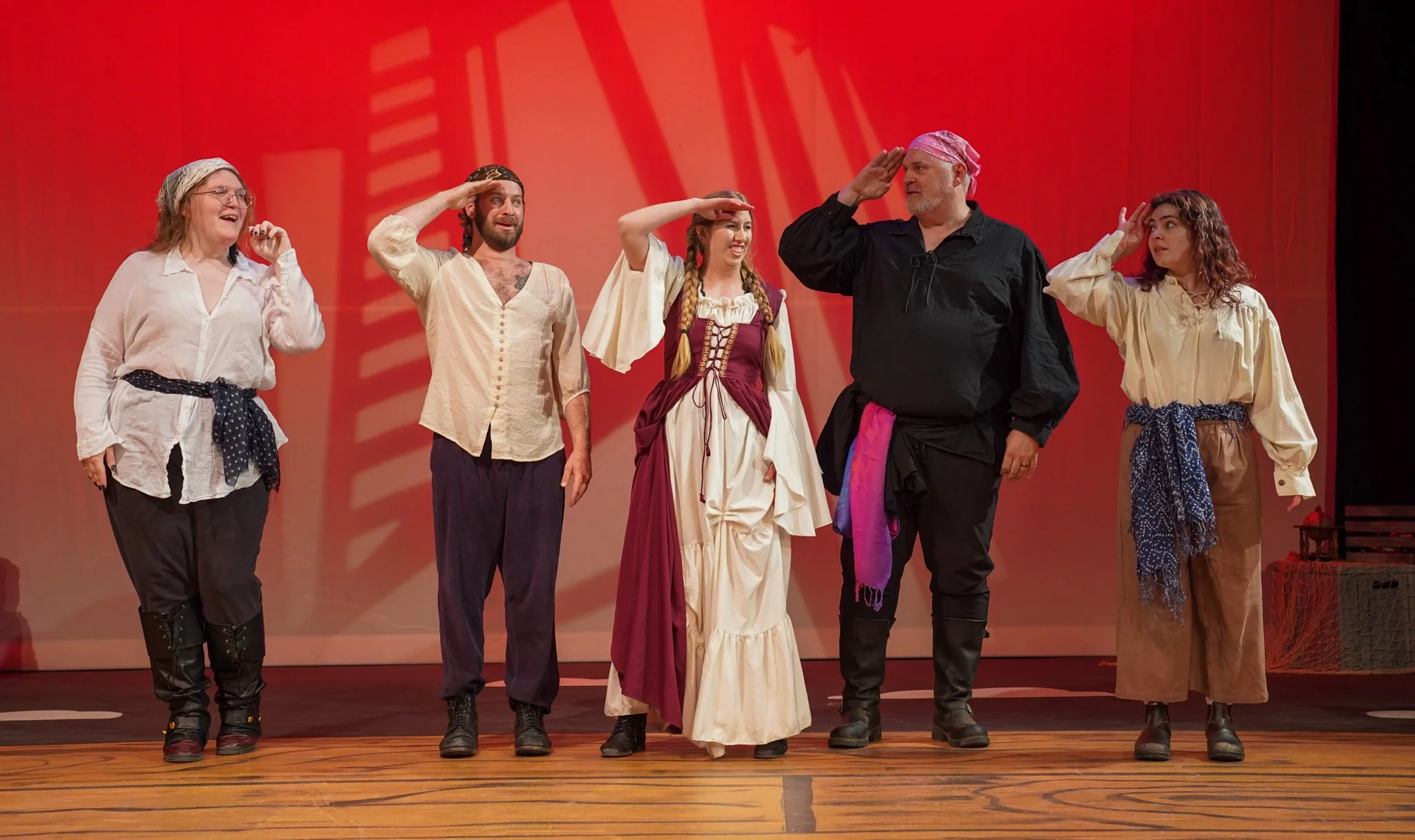 Group of six actors in costume on stage, saluting in a theater performance, with a red backdrop and wooden floor.