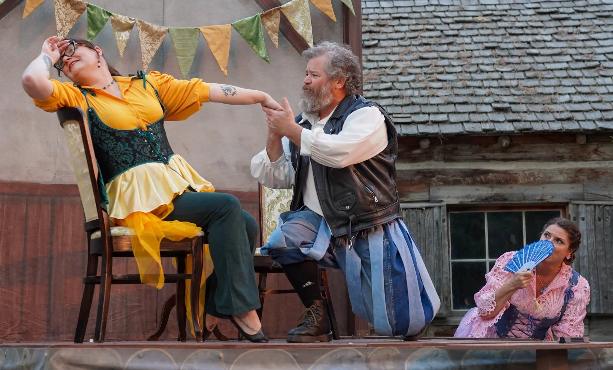 People performing a theatrical play outdoors, with one woman sitting on a chair wearing a yellow dress and glasses, another man kneeling in front wearing a vest and kilt, and a woman in a pink dress holding a blue fan nearby. Bunting decorations are 