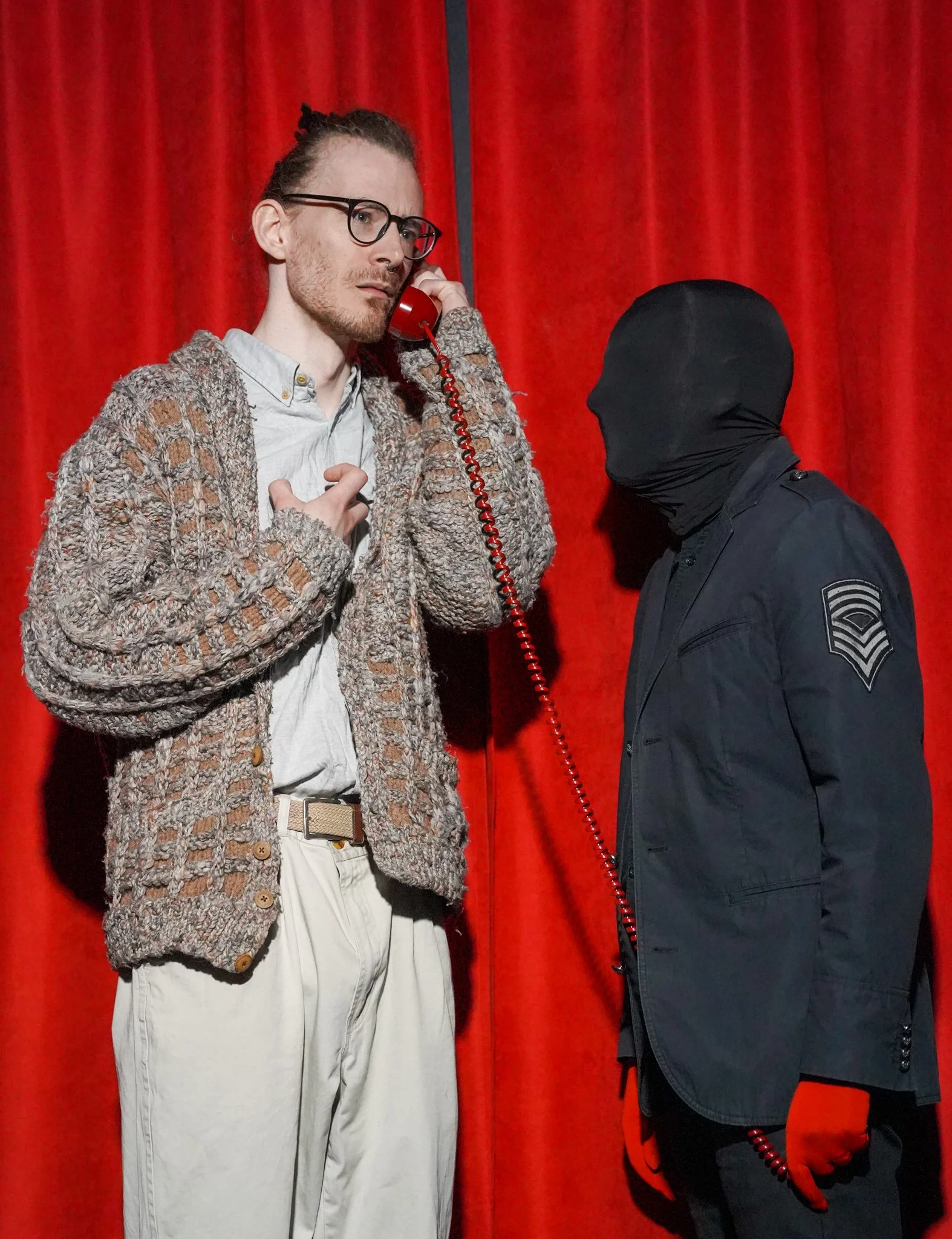 A man with glasses, a beard, and a pierced nose, dressed in vintage clothing, is on the phone with a police officer. The officer has a black balaclava and uniform, and they are standing in front of a red curtain.