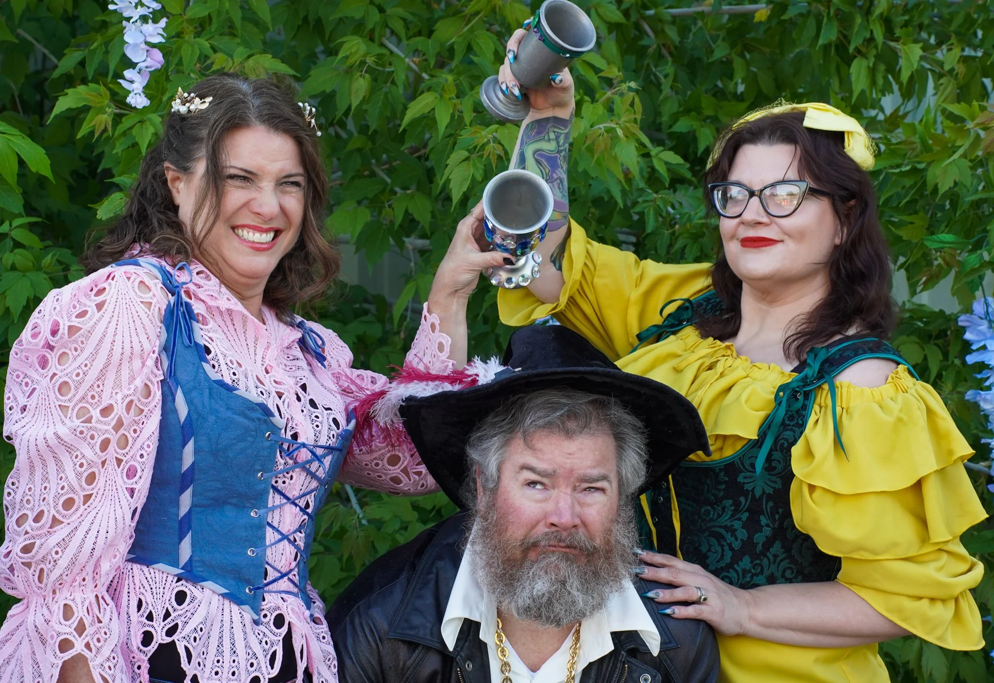 Group of people dressed as characters at a renaissance fair, with two women holding goblets, wearing colorful costumes, and a man in pirate attire in a leafy outdoor setting.