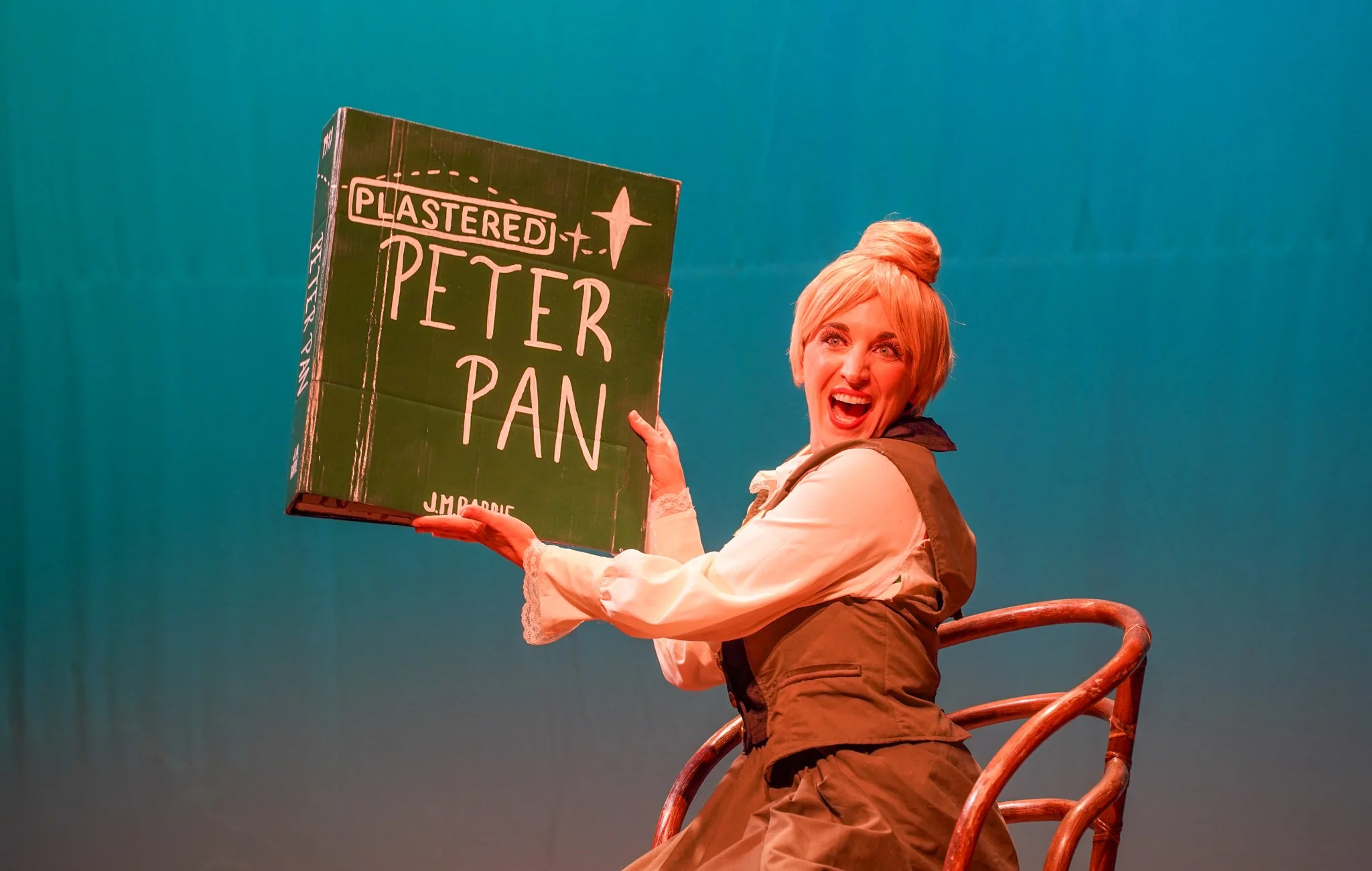 A woman with blonde hair styled in an updo, wearing period costume, sits on a chair with a wooden armrest, holding a green upper case sign that reads 'PLASTERED PETE R PAN' with a star symbol, against a teal background.