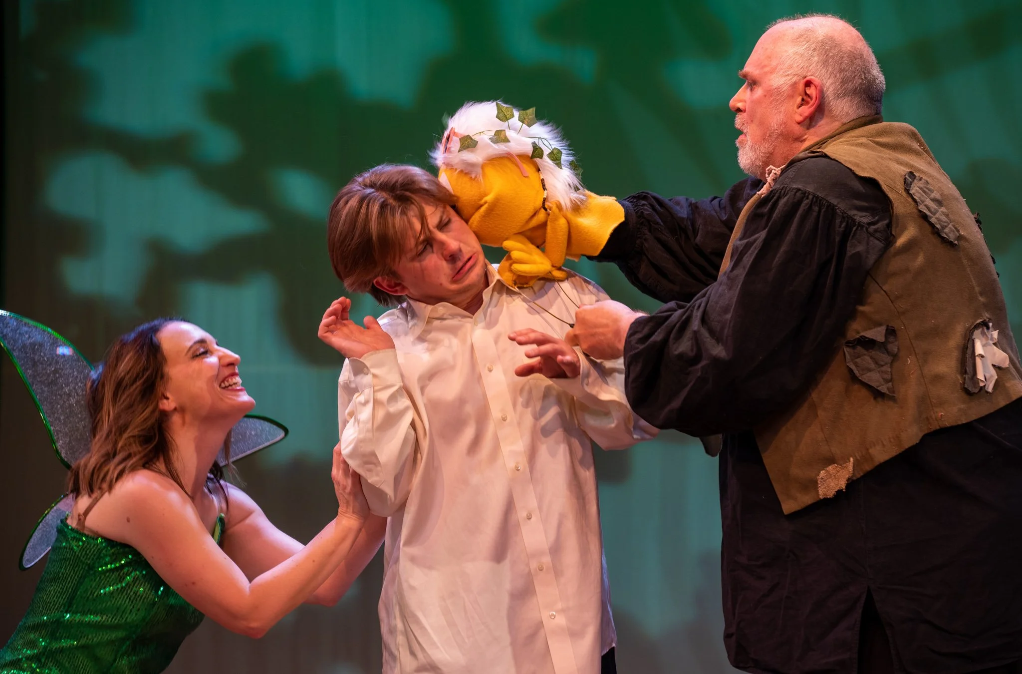 Four people dressed as fairies and a man with a puppet in a scene from a play or performance, with a green background.