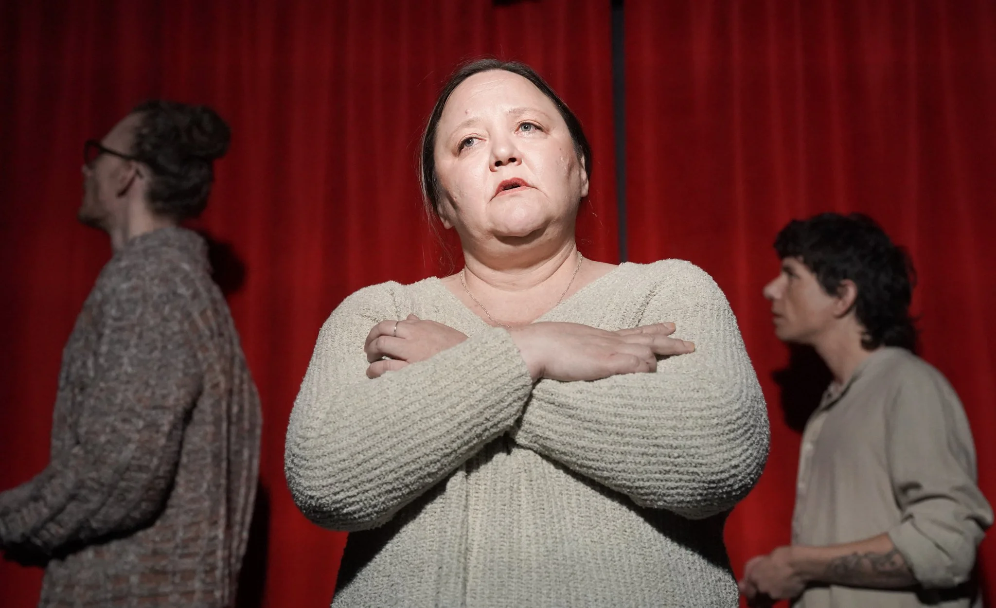 A woman with dark hair and pale skin, wearing a beige sweater, standing in front of a red curtain with her arms crossed over her chest, looking upward with a serious or contemplative expression. There are two other women in the background, one on eac
