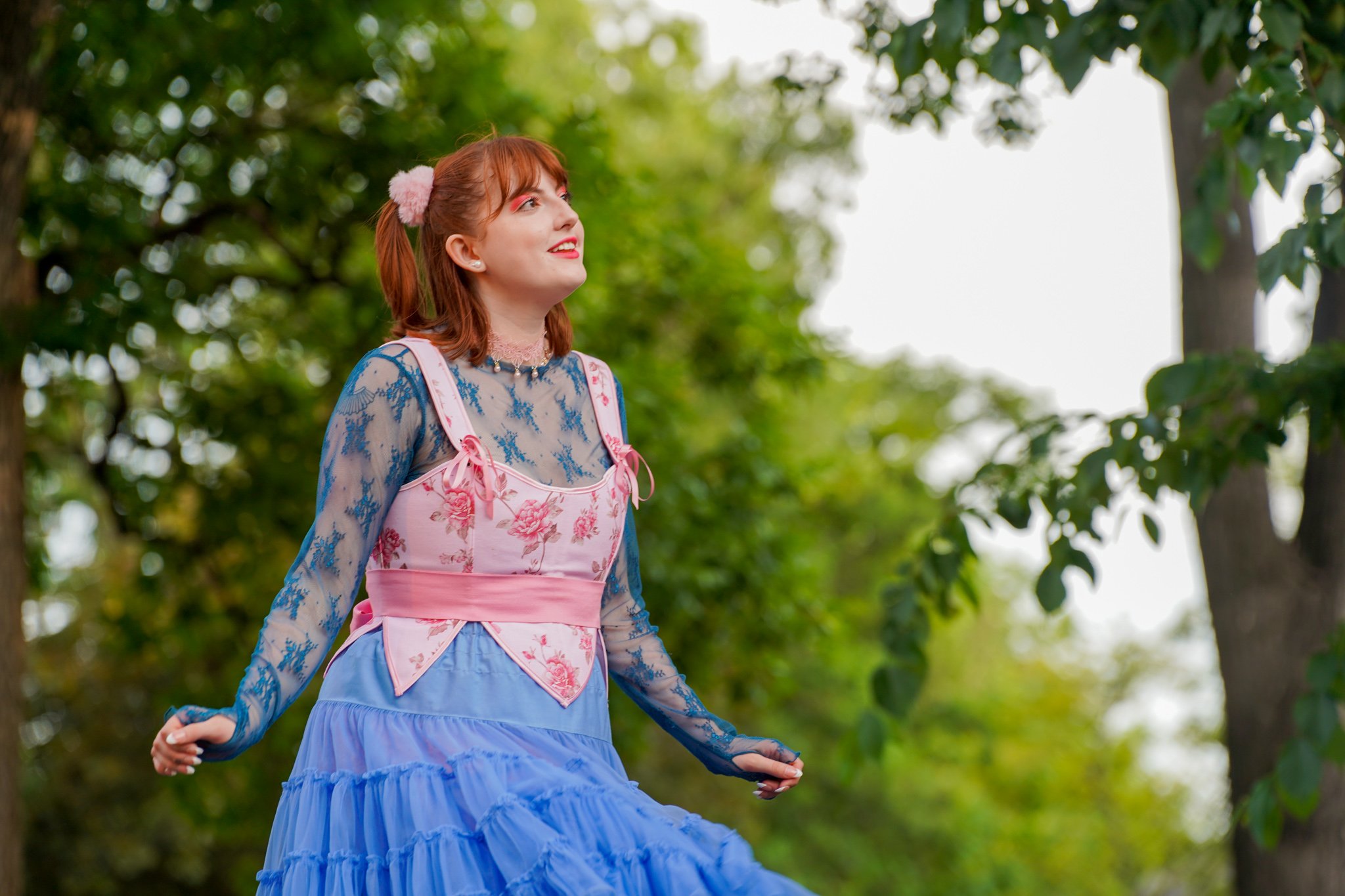 A young woman with red hair, dressed in a colorful, floral, gothic lolita-style outfit, is outdoors in a park surrounded by green trees, smiling and looking up.