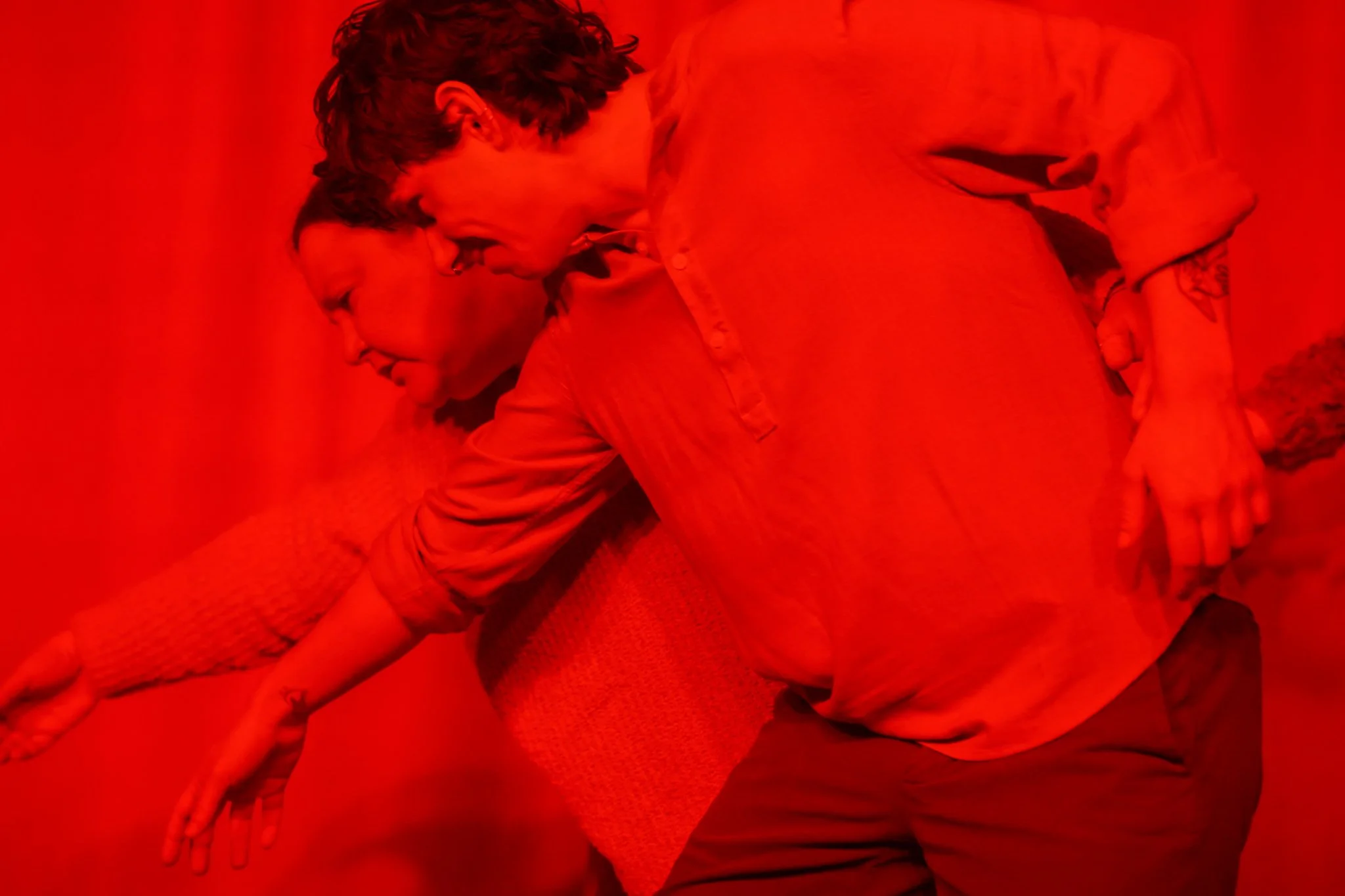 Two people are sitting on a couch against a red wall, facing each other with their foreheads touching and eyes closed, in a tender and intimate pose.