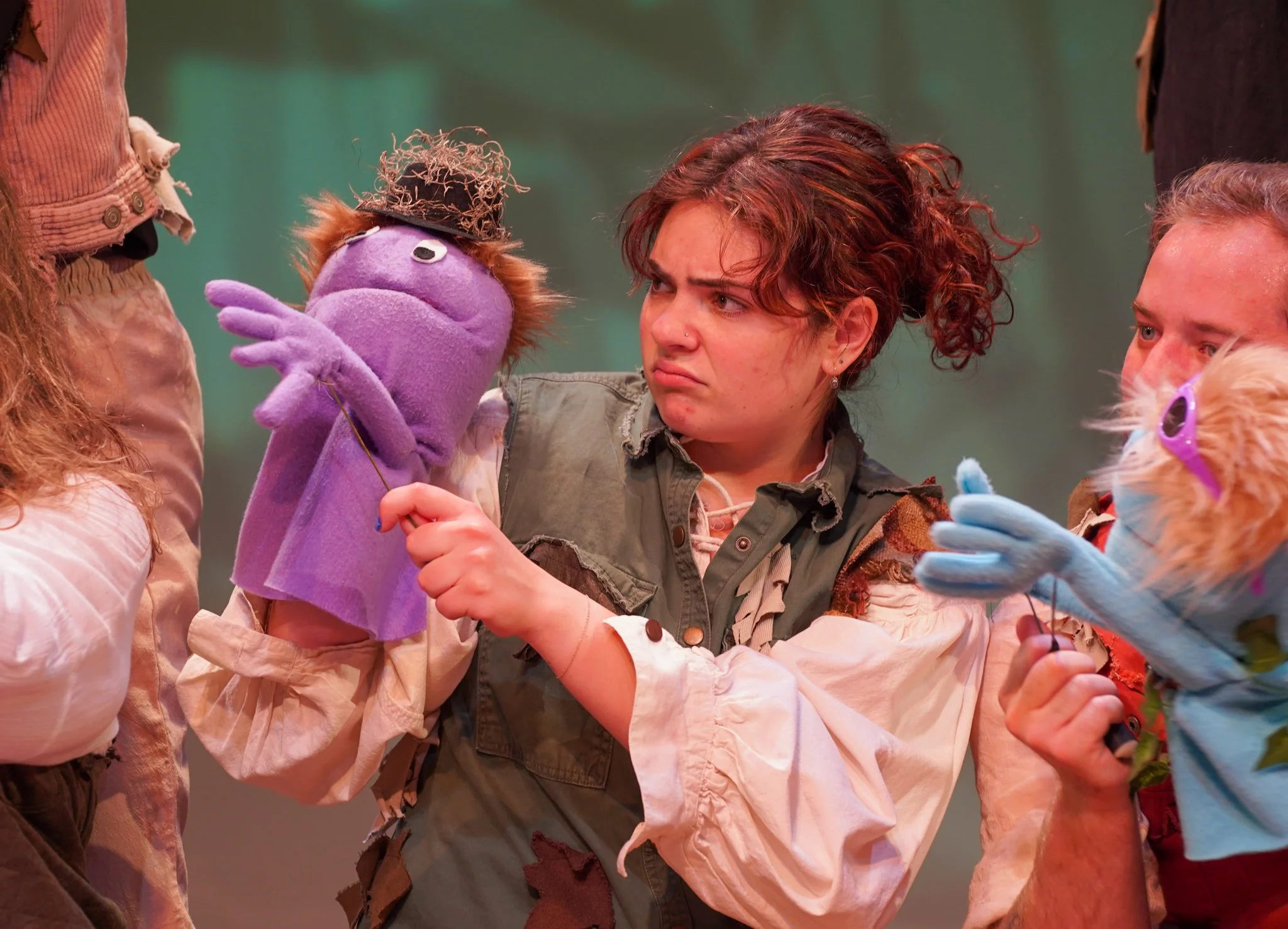 A woman with curly red hair holding a purple puppet with a black hat, engaging with another person whose face is partially visible, in an indoor setting.