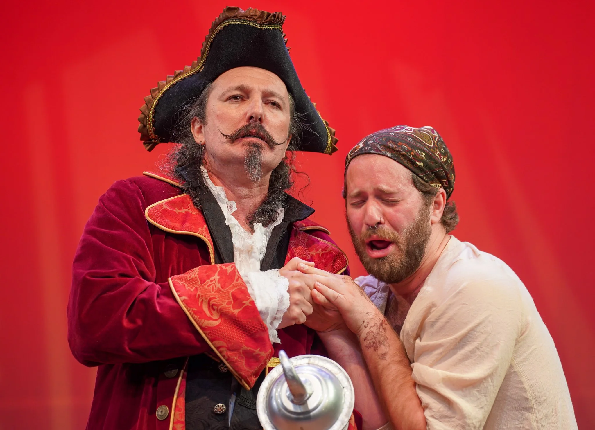 Two men dressed in theatrical costumes are holding hands in an emotional moment against a red background. One man is wearing a pirate or historical costume with a large black hat, red velvet jacket, and ruffled shirt. The other man is dressed casuall
