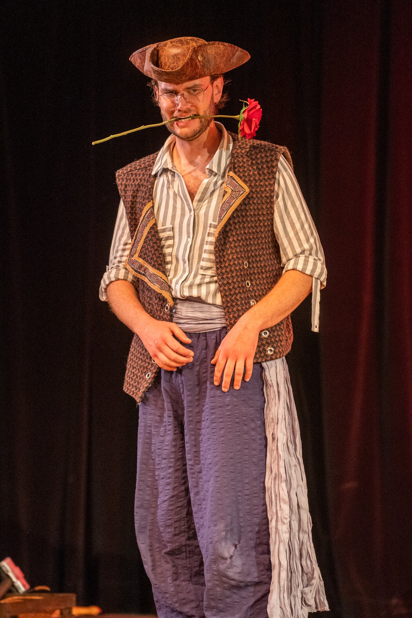 A man in costume stands against a black background, wearing a floppy hat, a vest, striped shirt, and baggy pants with a patch. He holds a red rose in his mouth and loosely clutches his hand near his waist, with facial expression showing a pout or fro
