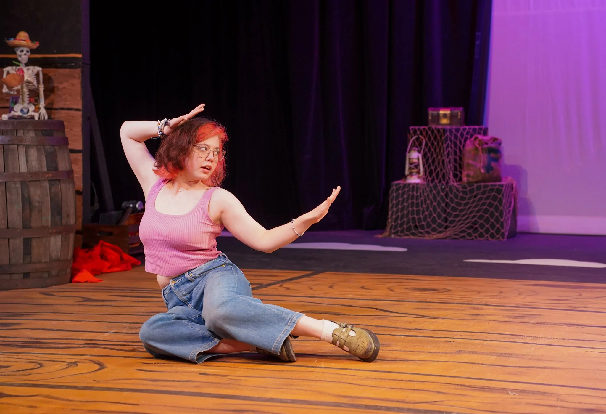 A woman with red and brown hair, glasses, wearing a pink tank top and blue jeans, is sitting on the wooden floor of a stage, with her legs crossed and hands raised in expressive gestures. The stage has a black curtain in the background, and there are decorated props including a skeleton with a sombrero, a barrel, and a netted chest with a pillow.