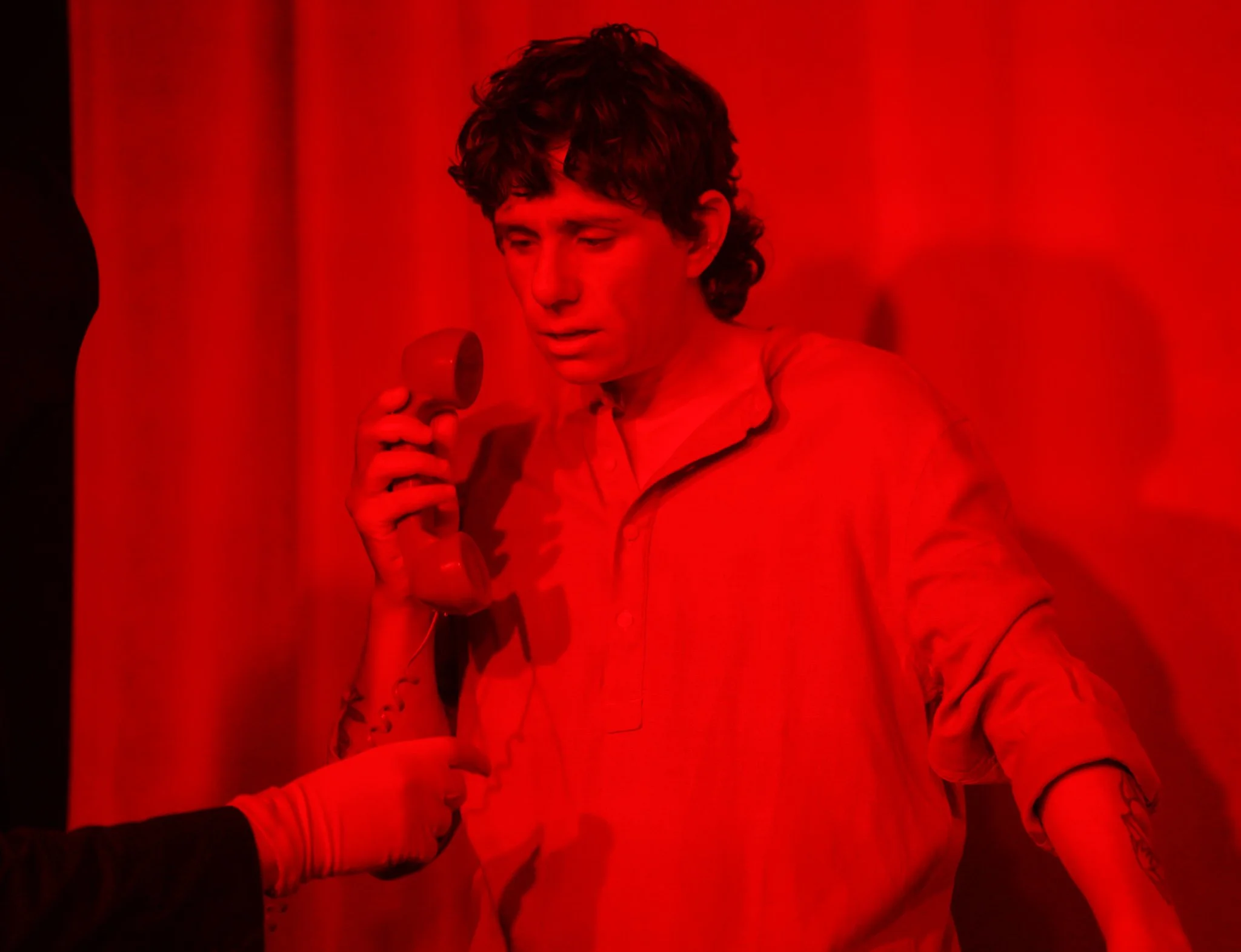 A young man with curly hair holding a red microphone in a red-lit setting.