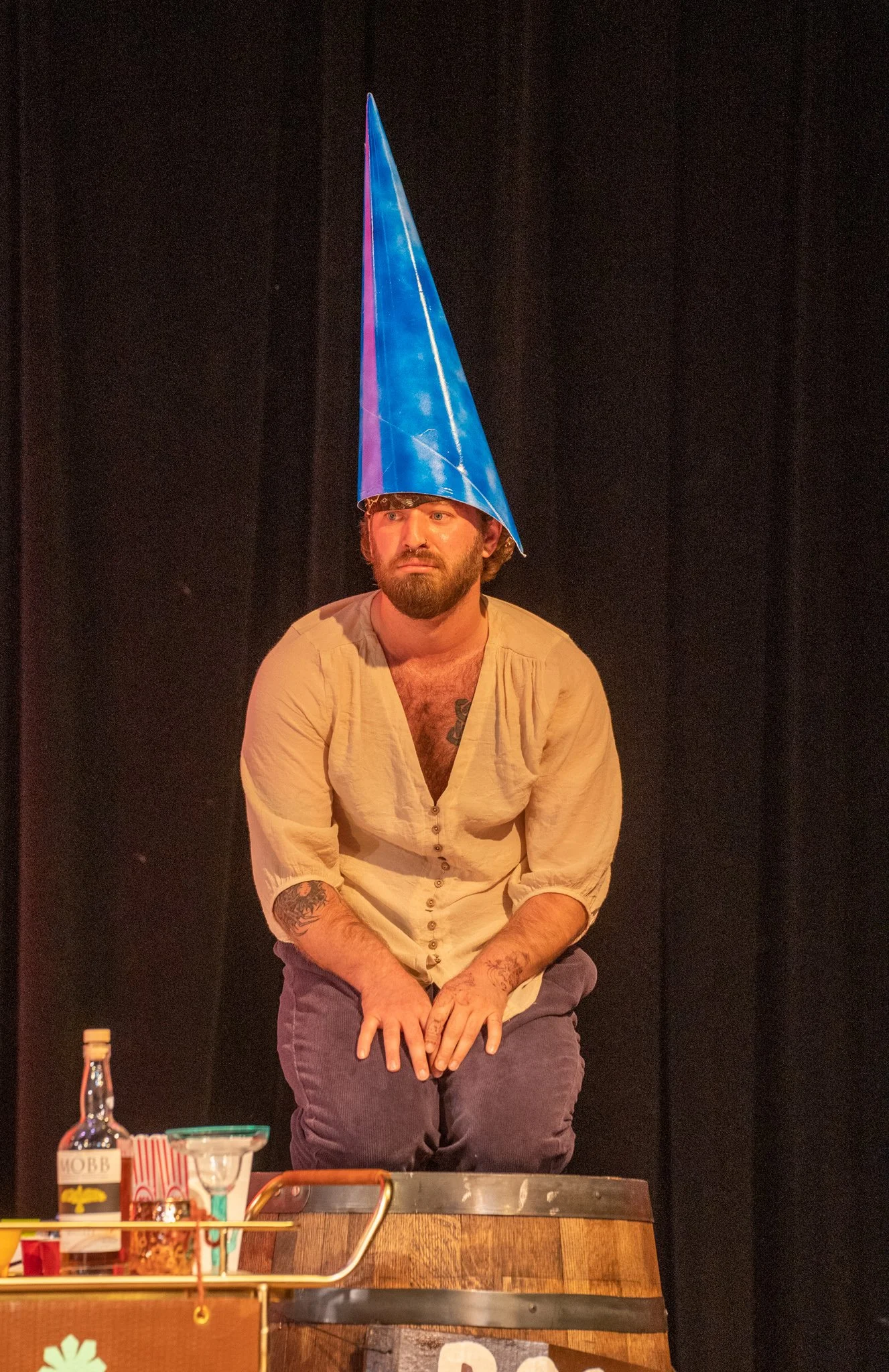 A man with a beard and tattoos on his arms, wearing a light-colored shirt and gray pants, sitting on a chair. He is wearing a tall, cone-shaped party hat with a blue and purple cloud design. There are various objects on a table in front of him, inclu