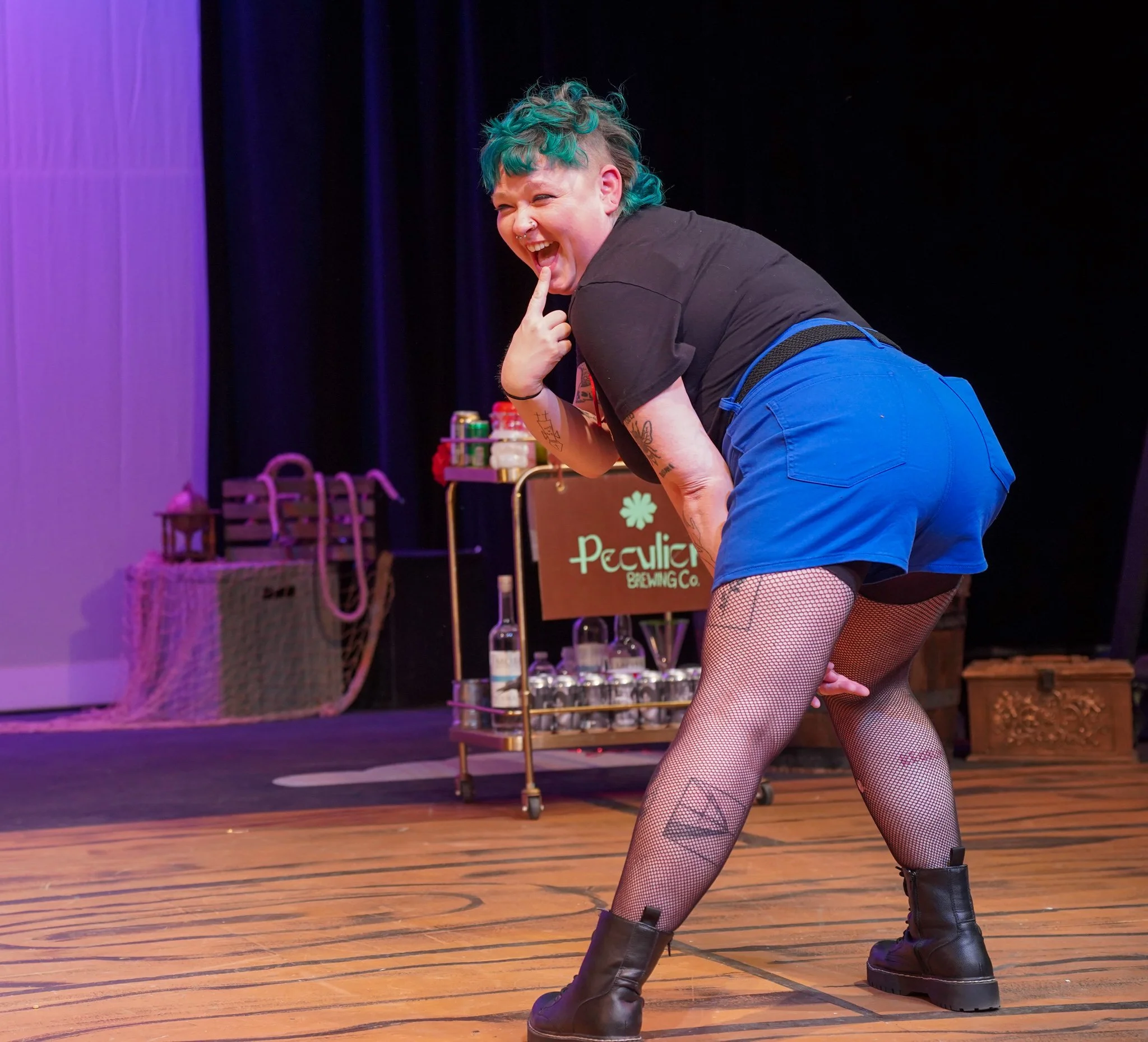 A person with teal curly hair, tattoos, fishnet stockings, and combat boots is posing on a stage with a big smile, holding a finger to their lips. The background shows a table with drinks and a sign for Peculier Brewing Co.