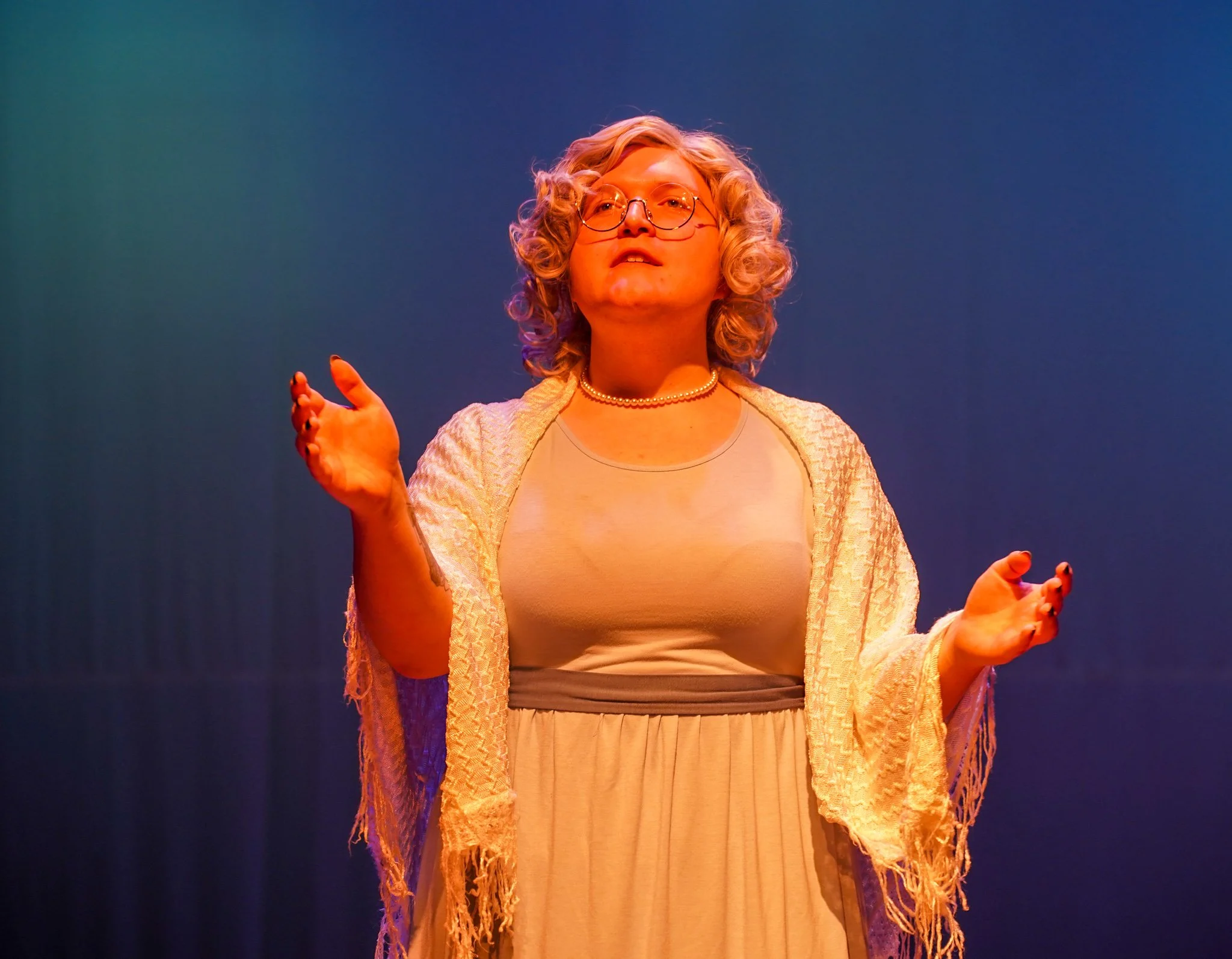 Woman with curly blonde hair and glasses, wearing a beige dress and pearl necklace, speaking on stage with blue and orange lighting.