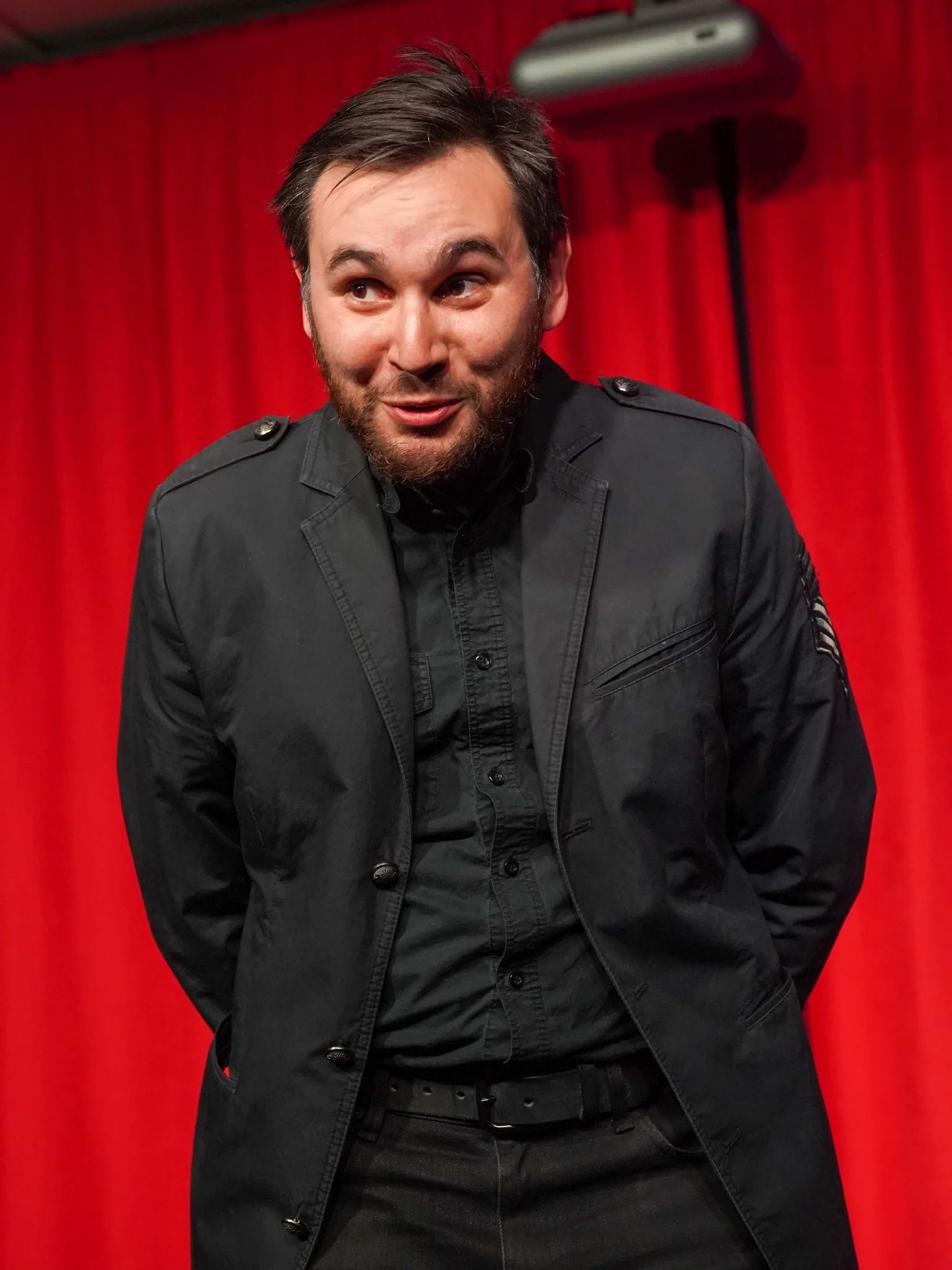 A man sits in front of a red curtain, wearing a black jacket and black pants, with a beard and short hair, looking slightly to the side and smiling.