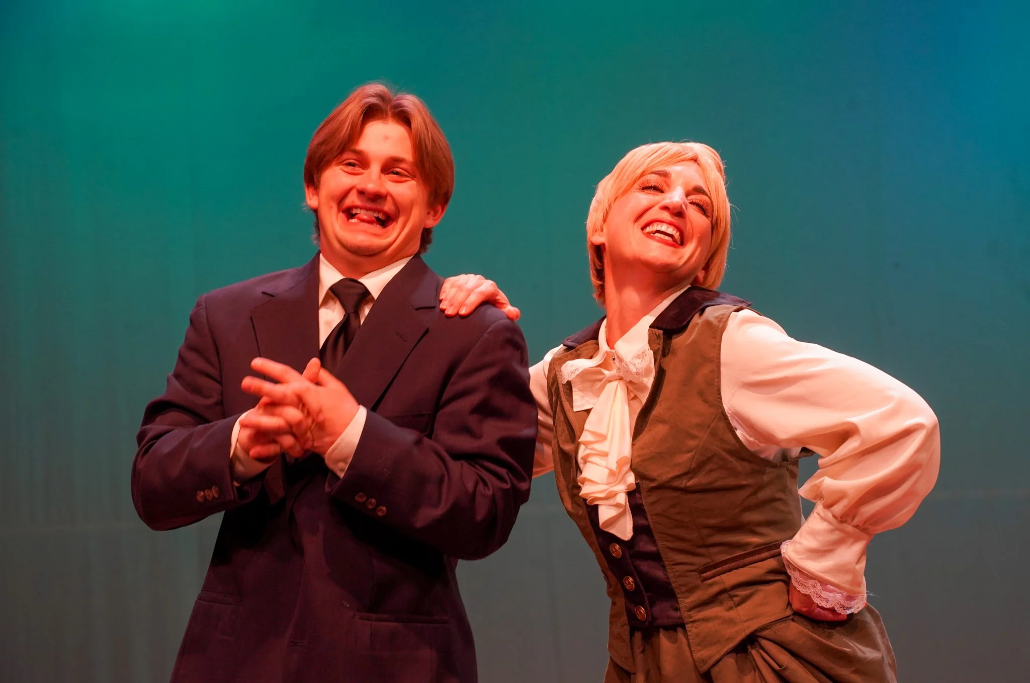 Two performers on stage, a young man in a black suit and a woman in a period costume, both smiling and posing playfully.