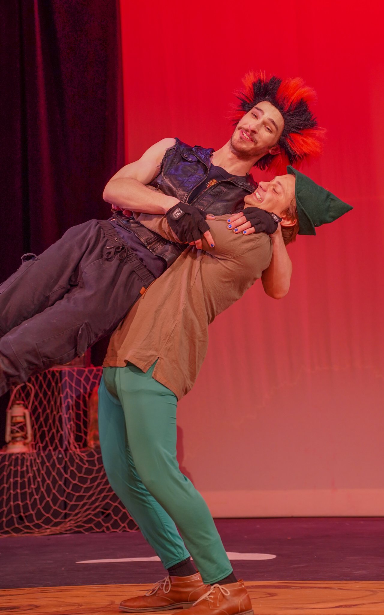 Two men in theatrical costumes dancing on stage against red background. One man has red and black spiky hair, wearing a black leather vest and tan gloves. The other man wears a green hat and green pants.