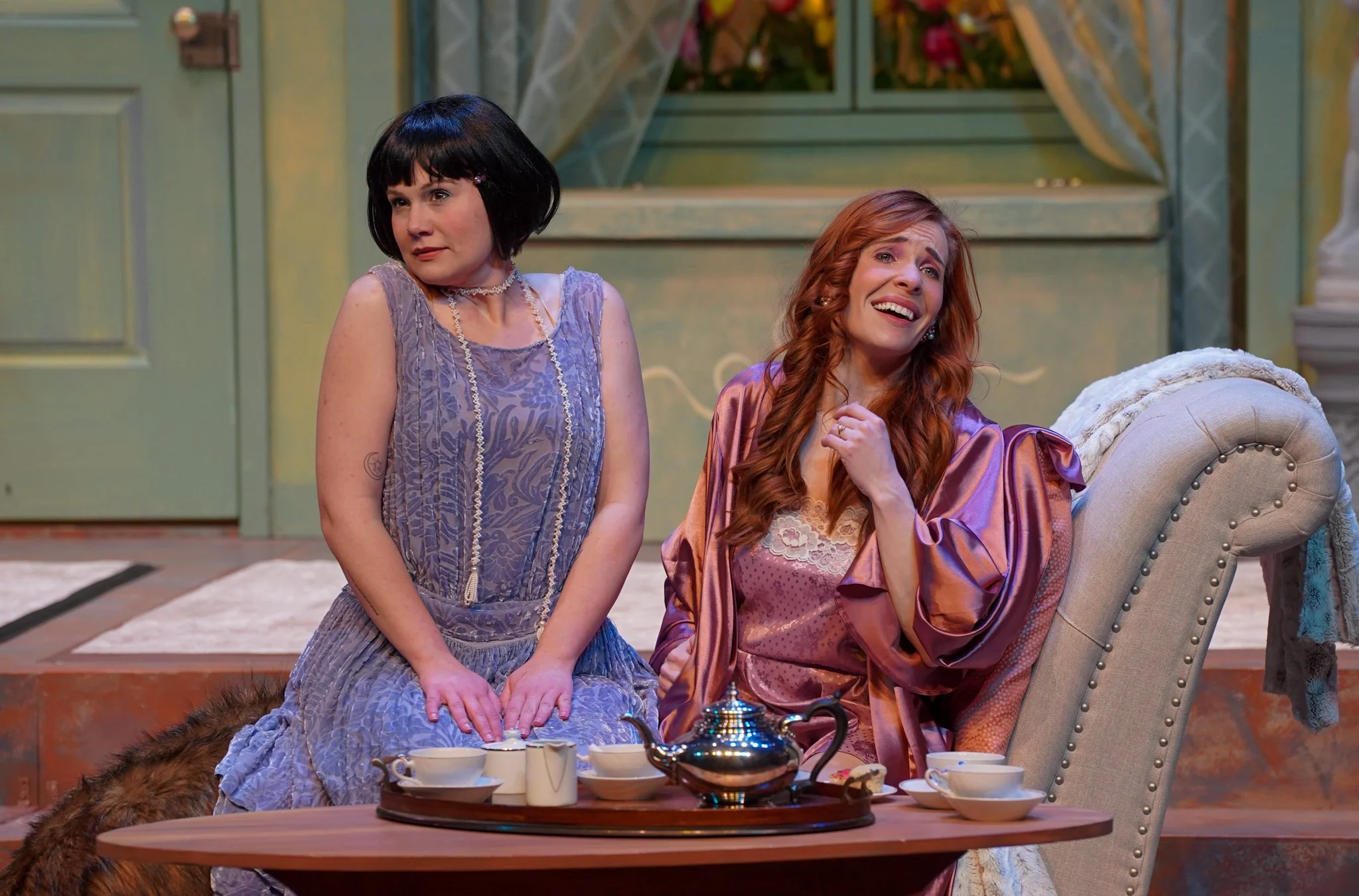 Two women sitting at a tea table, one with black hair in a bob wearing a lavender dress, and the other with red hair in a silk robe, both appearing emotional; the scene resembles a theatrical play set with a pastel-colored background and a table with