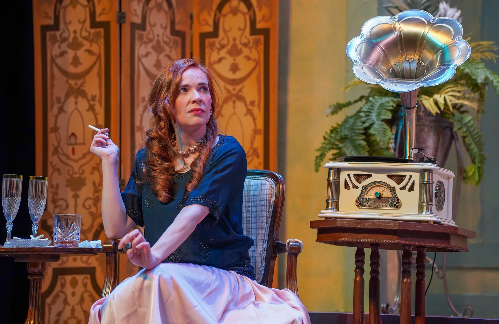 A woman with red hair wearing a blue top and pearl necklace sits at a table with empty glasses, holding a cigarette in her right hand and looking thoughtfully. There is a vintage record player on a small table behind her, and decorative wallpaper and