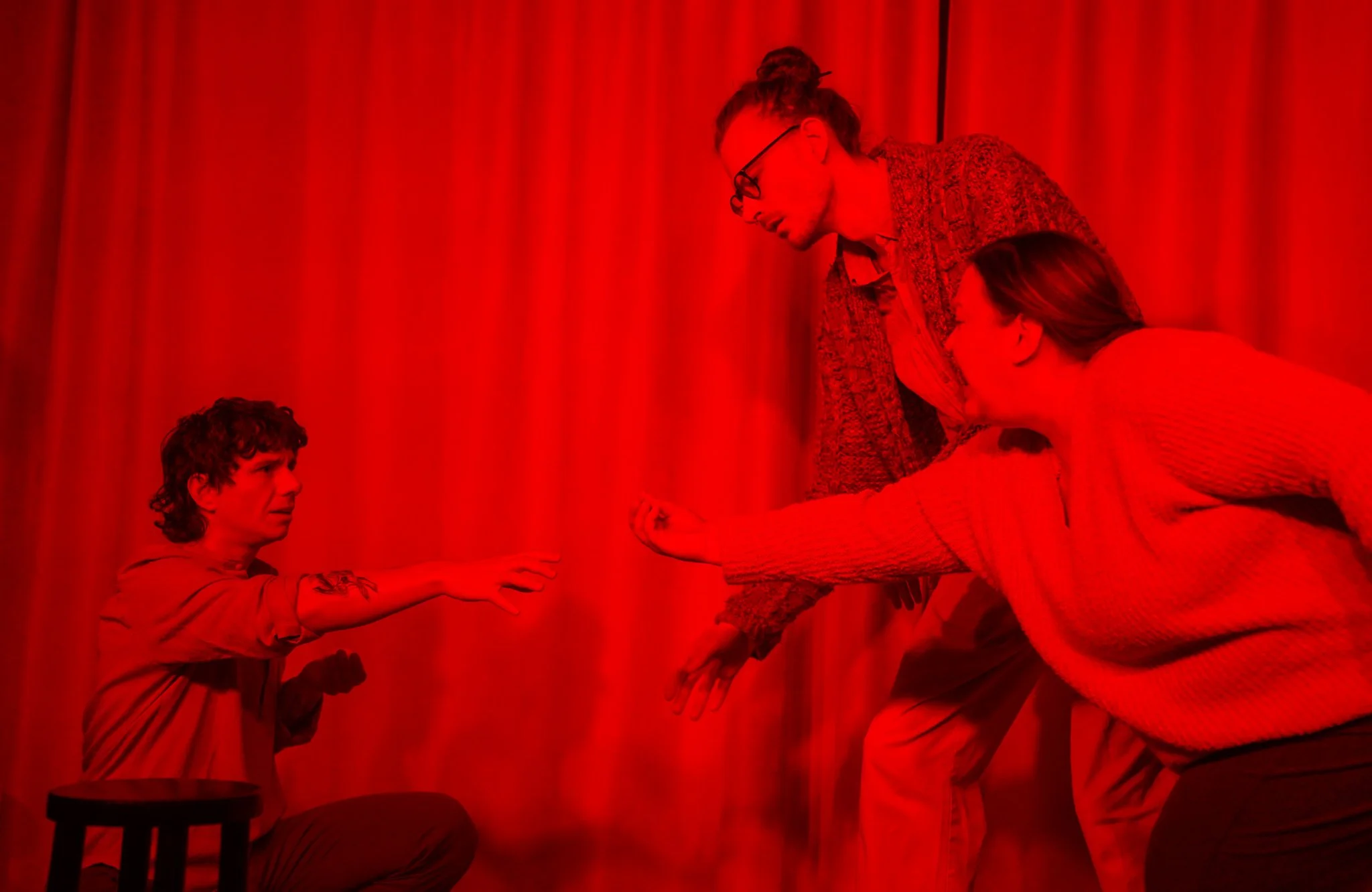 A person is lying on their side on a bed, reaching out to a young boy who is sitting and extending his hand towards the person. Another person is standing, leaning over, assisting or engaging with the boy. The background consists of a red curtain, an