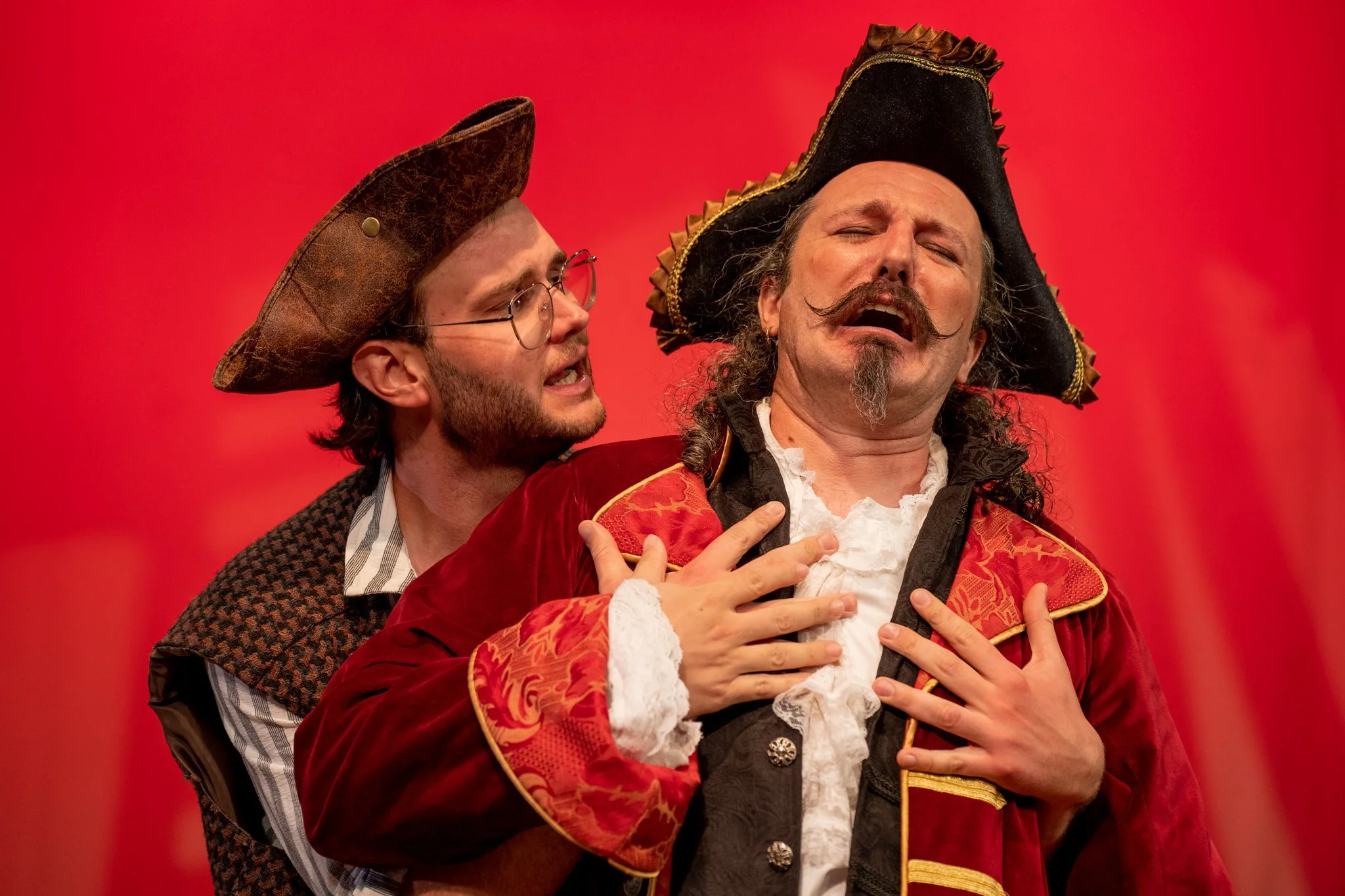 Two men perform in costumes resembling pirates, with one dressed in a red and gold coat and the other in a brown coat and glasses, against a red background.